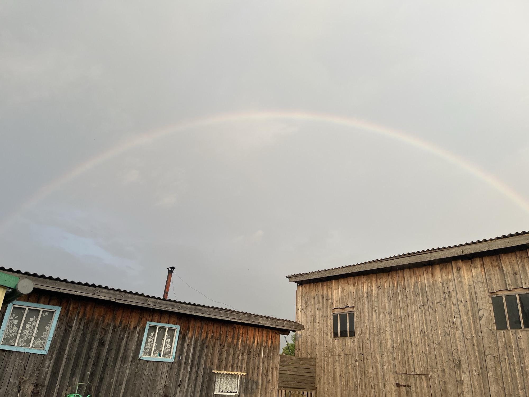 In the city of Bolshoi Gondyr, Russia, a faint rainbow arches across a cloudy sky above two wooden structures. The structure on the left, with a corrugated roof, features two windows with light blue frames, both covered with white lace curtains. A metal chimney extends from its roof. To the right, a larger, taller wooden building with a similar corrugated roof has two windows, one near the top and another lower down. The scene is devoid of any people or animals. The environment appears to be rural, with the weathered wood of the buildings suggesting an outdoor setting.