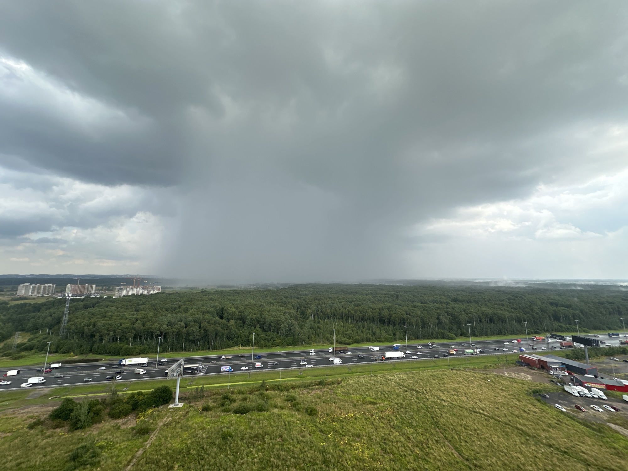 An elevated, wide-angle shot captures a dense forest bordering a multi-lane highway in Imeni Sverdlova, Russia. Dark, heavy storm clouds dominate the sky, with a distinct rain shower visible in the distance, creating a hazy curtain over the landscape. The highway is active with a stream of vehicles, including cars and trucks. On the left side of the highway, residential buildings under construction are visible, suggesting urban development encroaching on the natural environment. The right side of the highway features industrial buildings and a parking lot filled with various vehicles, including trucks and vans. A grassy field with scattered yellow wildflowers extends from the foreground towards the highway, adding a touch of natural color to the otherwise somber scene. The overall atmosphere is one of impending or ongoing bad weather contrasting with the everyday activity of traffic and construction.