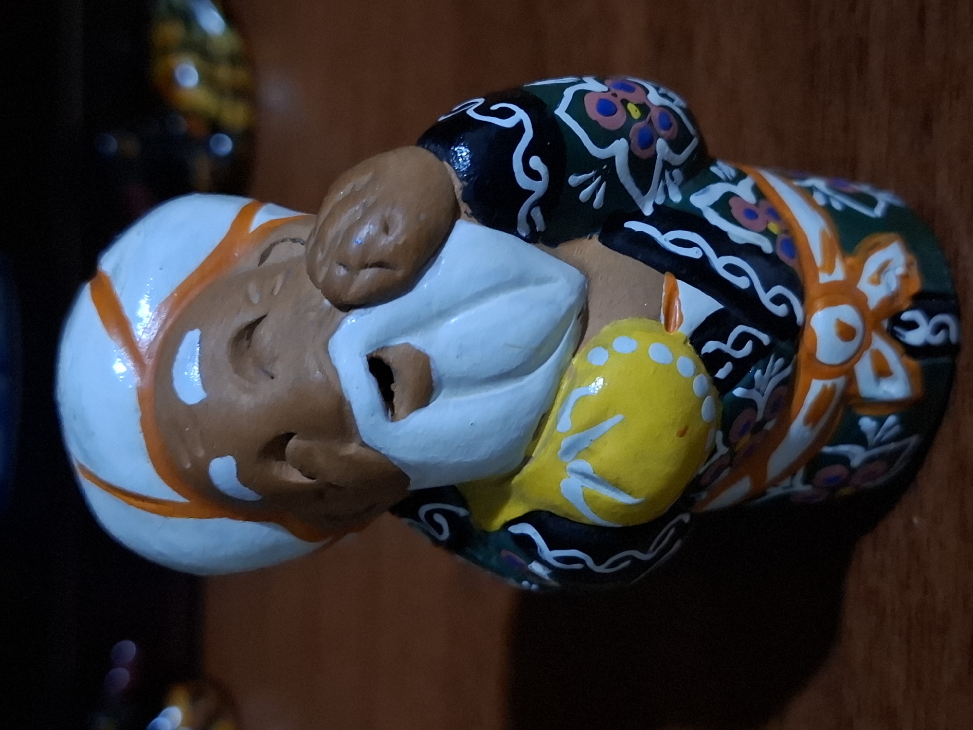 A small, brightly colored ceramic figurine of a laughing old man wearing a turban and traditional clothing stands on a wooden surface. The figurine has a brown face with a white beard and is holding a yellow bag. The background is dark and out of focus, with some blurred lights visible. There is no indication of the city of Almaty, Kazakhstan, or any specific location within it.
