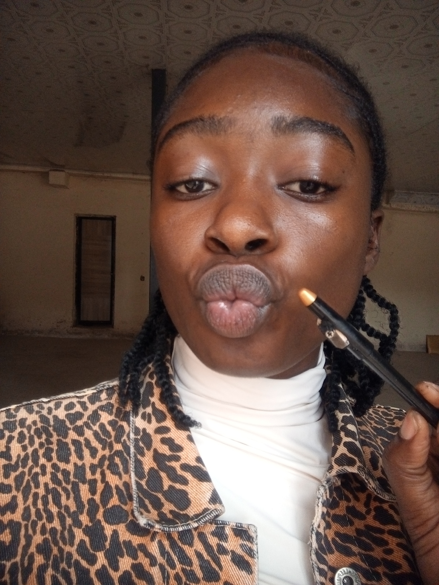 A close-up selfie captures a young Black woman with braided hair against a blurred background of an indoor space. She is wearing a white turtleneck and a leopard-print jacket. Her lips are pursed, and she is holding a golden cosmetic pencil near her mouth. The background suggests an unfinished or sparsely furnished room with a door visible on the left wall and a textured ceiling.