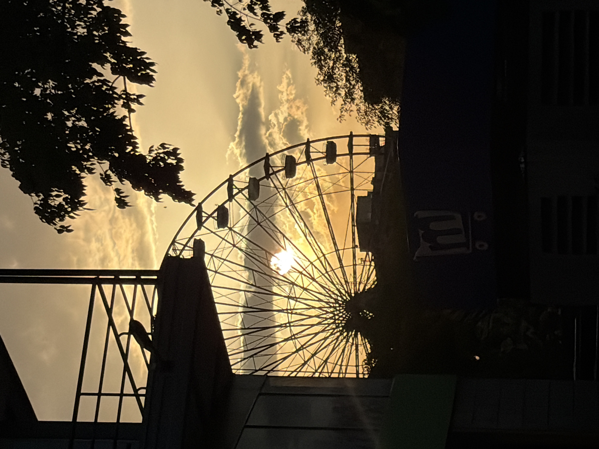 The image shows a Ferris wheel silhouetted against a sunset sky. The sun appears to be setting directly behind the center of the wheel. Trees are visible on either side of the Ferris wheel. Below, part of a building and fence are visible. The location is likely Ciudad Guayana, Venezuela.