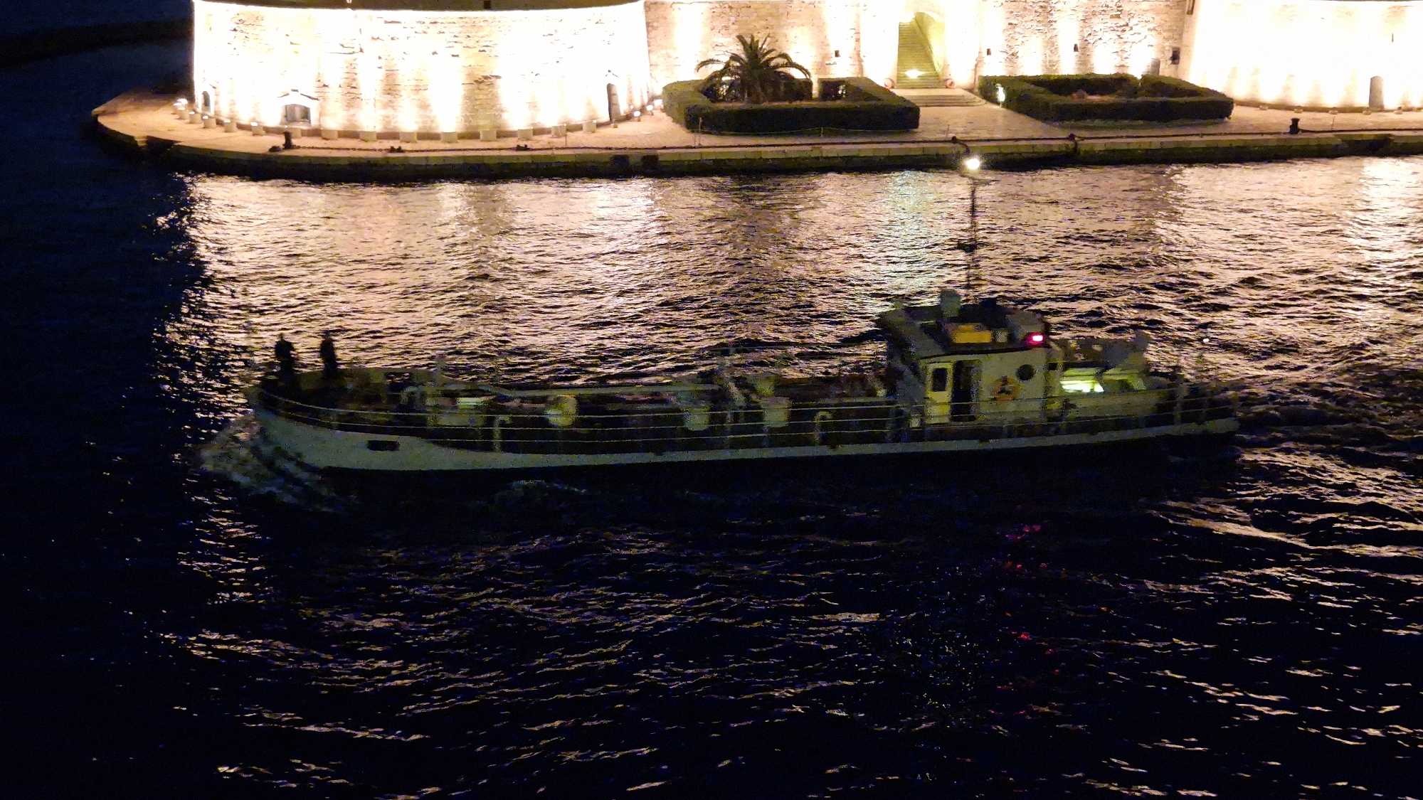 A nighttime aerial shot shows a boat on the dark water of the Mar Piccolo in Taranto, Italy. The boat, which appears to be a small cargo ship or barge, is white with yellow accents and has a cabin structure towards the stern. It is illuminated by interior lights and a red light at the top of its mast. Two figures are visible on the deck of the boat. The water is choppy, reflecting the lights from the nearby shore. In the background, a large, circular, fortified structure, likely part of the Aragonese Castle in Taranto, is brightly illuminated with warm lights. The walls of the structure are stone, with vertical elements and arches. There is landscaping on the ramparts of the fortification, including a palm tree and manicured bushes. A walkway or quay separates the water from the fortification.