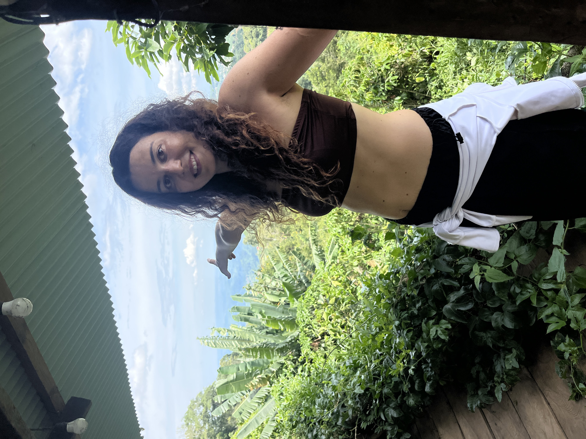 A woman with curly brown hair stands on a wooden deck, pointing to the right with her left hand. She wears a brown tube top, black pants, and a white jacket tied around her waist. The background shows lush green vegetation, including banana trees, and a hazy landscape with distant hills under a cloudy sky. Two light bulbs hang from the corrugated metal ceiling of an overhang. The location is described as the city of Son Dien, Vietnam, though this specific location within Son Dien is not discernable from the image.