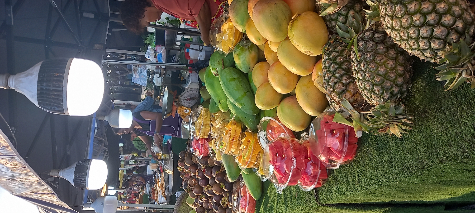 This image shows a fruit stand in Thong Sala, Thailand. The vendor and customer are obscured by a display of mangos, papayas, pineapples, mangosteens, and pre-cut fruit in plastic containers. A weighing scale is visible on the countertop. The stand is illuminated by modern LED lights. The artificial turf ground cover is visible in the lower right.