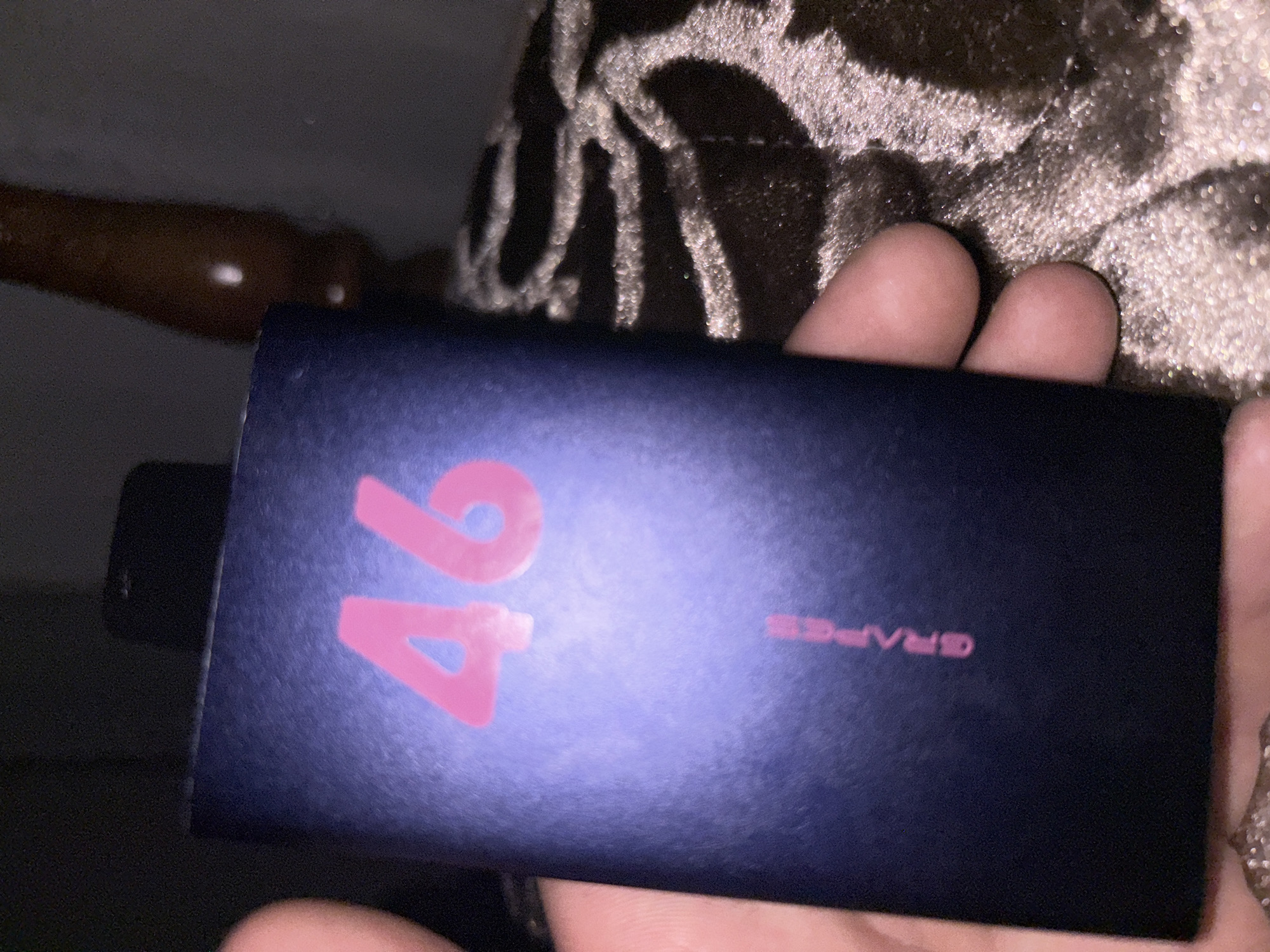 The image shows a close-up of a rectangular, dark blue object, possibly a vape device or a small power bank, held in a person's hand. The object has the number "46" printed in bright pink on its surface, and below that, the word "GRAPES" is also printed in pink. The background is dark and appears to be an indoor setting, with a textured, possibly upholstered surface visible. There is no indication of the location being in Paraul, Russia, or any other specific city.
