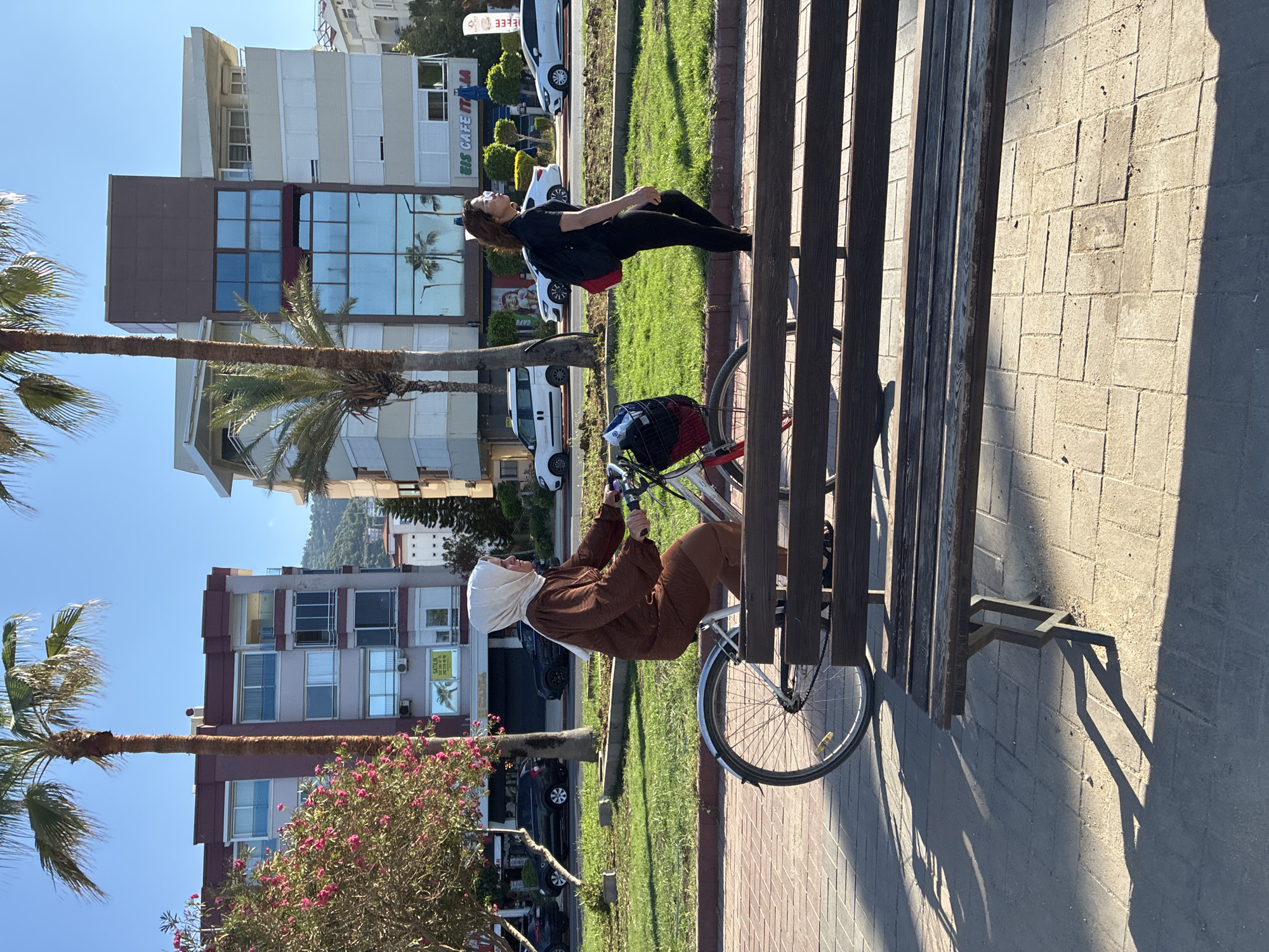This media file captures a sunlit outdoor scene in Alanya, Turkey, likely a promenade or park area adjacent to a street with buildings. The bright sunlight casts distinct shadows, suggesting late morning or afternoon on a clear day.
In the foreground, a sturdy wooden slatted bench runs parallel to the paved walkway made of light-colored interlocking pavers. Two women are present:
One woman, wearing a black top, black leggings, and sunglasses, is seen relaxing on a grassy patch beside the path. She has a red bag positioned nearby.
Closer to the bench, another woman, dressed in a brown long-sleeved outfit and a light-colored head covering (hijab), is seated on a bicycle. She appears to be adjusting something on the handlebars or within the front basket of the bicycle, which has a white frame and a red lining in its basket.
The background features several multi-story buildings, some with reflective windows showing reflections of palm trees. One building clearly displays partial text: "EIS CAFE IT" and "M", likely indicating "Eis Cafe Italia". Another building further back has a partially visible "SATILIK" sign (Turkish for "for sale"). Palm trees are prominent throughout the scene, interspersed with green grass and a vibrant bush with pink/red flowers, reinforcing the warm climate. Several white cars are parked along the street in the distance. The overall impression is one of a casual, sunny day in an urban coastal environment.