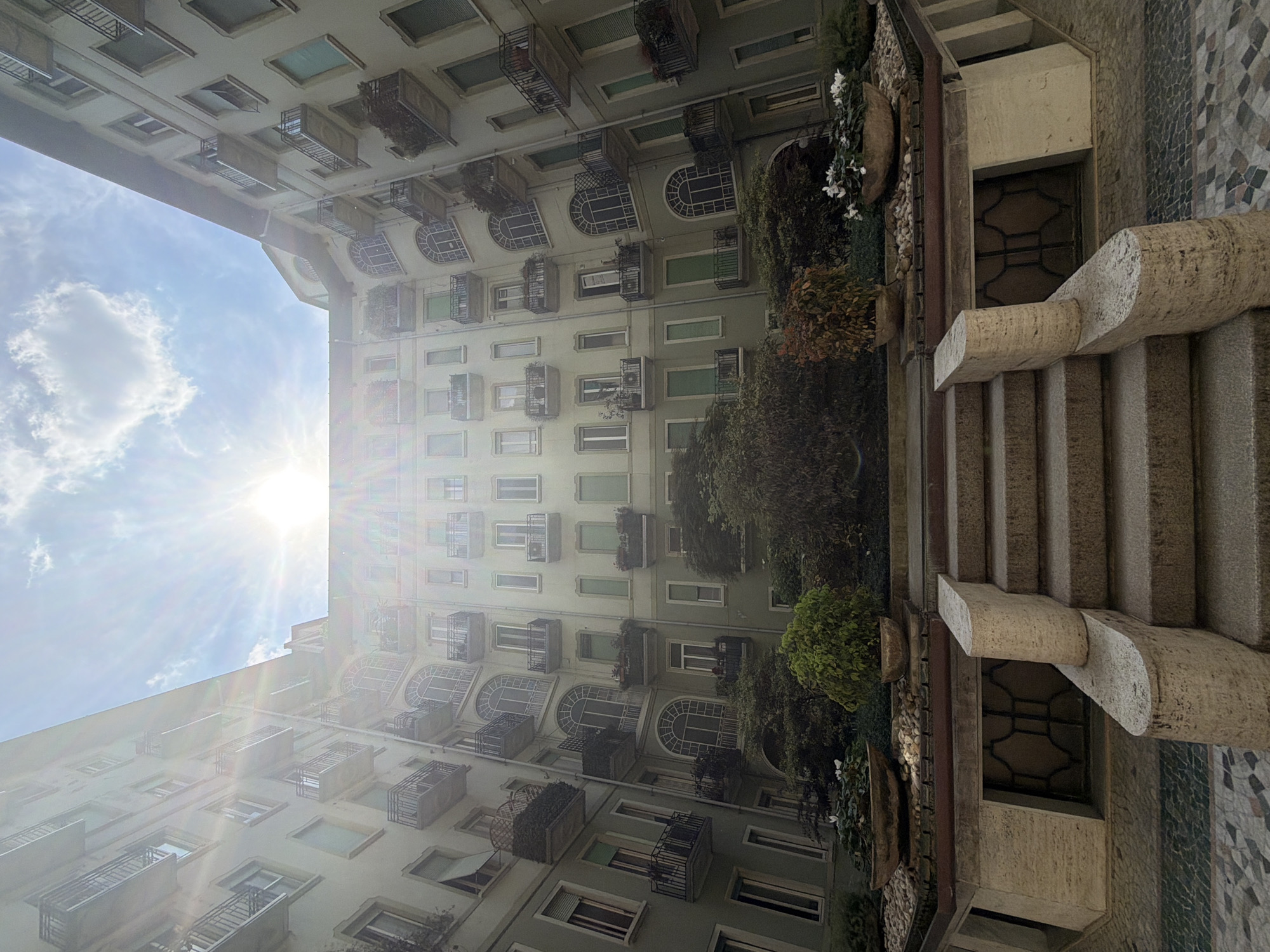 The image presents an upward-looking, slightly rotated perspective into a sun-drenched urban courtyard, consistent with the architectural style found in Milan, Italy. Multi-story buildings with light-colored facades form the dominant elements, featuring numerous windows and balconies. Many windows are rectangular, while others display elegant arched tops, some fitted with green shutters. Small iron balconies and window boxes filled with green plants adorn the facade, creating a vibrant contrast against the building.
The bright blue sky, with scattered white clouds, fills the upper left portion of the frame. A brilliant sun shines intensely from behind the building, creating strong sunbeams and significant lens flare across the scene, indicating clear daylight.
In the bottom right, a sturdy stone staircase with thick, textured balustrades descends into the courtyard. Adjacent to the stairs, a decorative mosaic or tiled wall with geometric patterns is visible. Lush green trees and bushes grow along the base of the buildings and near the staircase, with clusters of white flowers adding delicate detail. A window with a decorative metal grate is partially visible beneath the stair structure. No discernible human activity or text is visible.