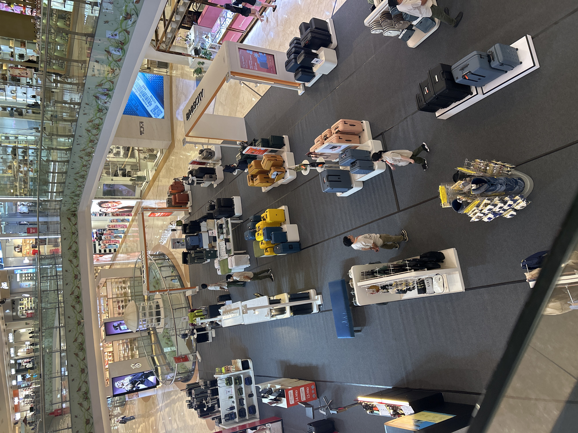 The image captures an overhead view of a luggage display within a large, modern shopping mall. The mall appears to be multi-level with a central atrium. The primary focus is on a section of the ground floor dedicated to a luggage sale, possibly a pop-up store, identified by signage reading "BIGESCITY". Numerous suitcases in various sizes, colors (black, blue, yellow, pink, tan, and green), and brands are neatly arranged on display stands and platforms. Some shoppers are present, casually browsing the merchandise. In the background, other retail stores and escalators are visible, indicating the bustling commercial environment of the mall. Based on the "BIGESCITY" branding and common mall layouts in Indonesia, this scene is likely located in the Trans Studio Mall in Makassar, Indonesia.
