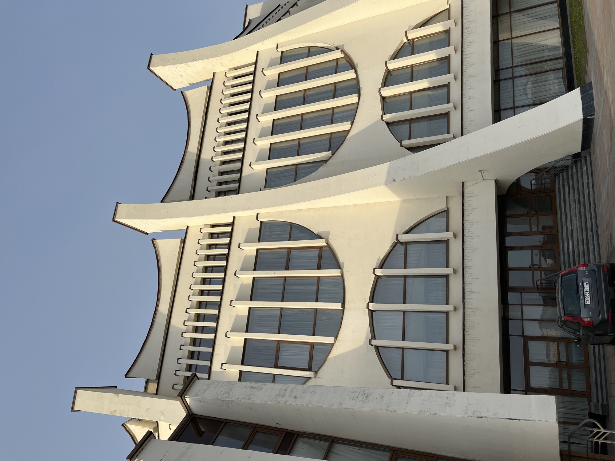 The image shows an architectural exterior in Unknown, Unknown. The building is white with distinctive curved elements and circular windows. A small car is parked to the right of the building entrance. The sky is clear and light blue.