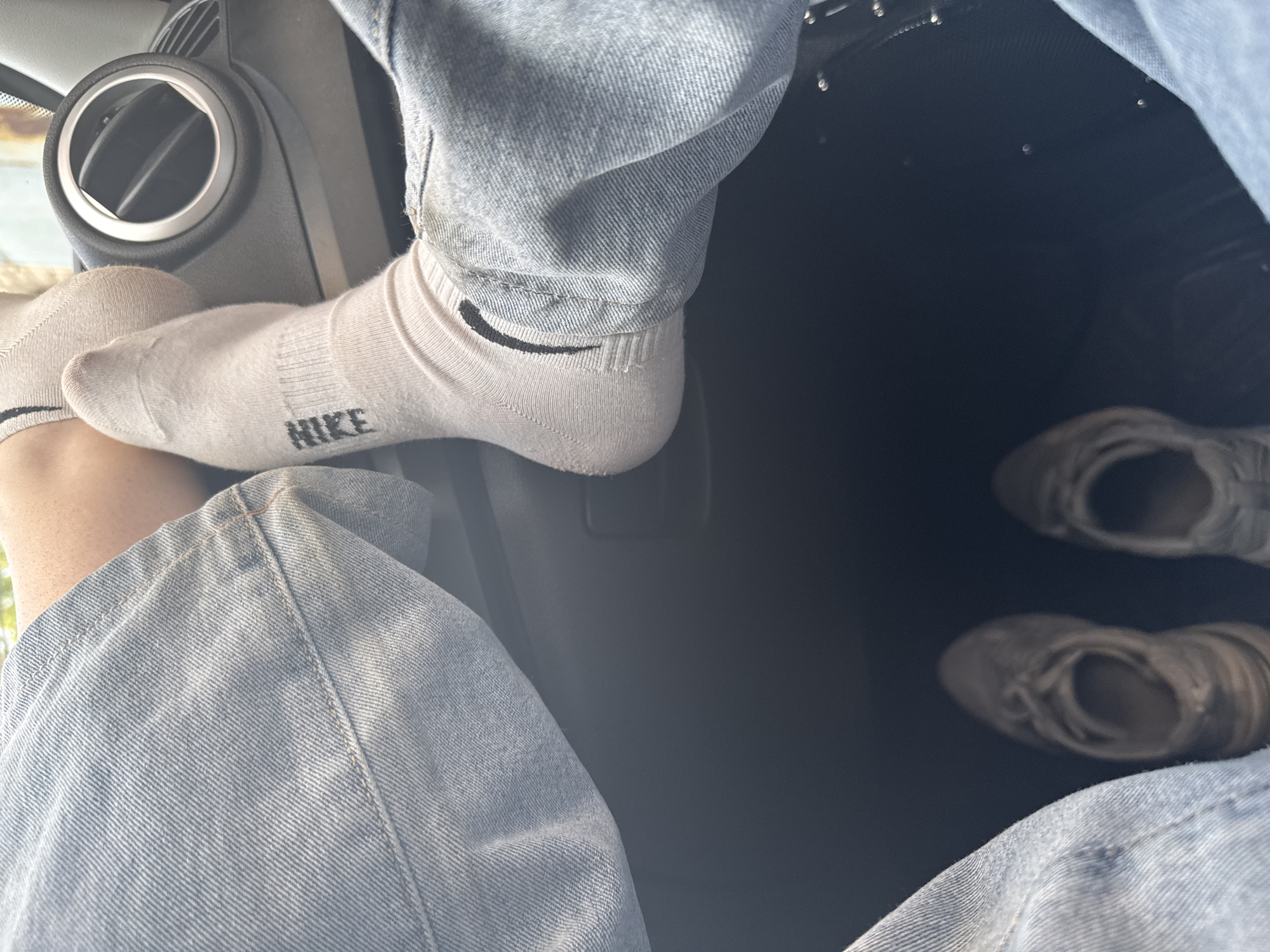 The image presents a high-angle, close-up view of a person's lower body within a car interior, likely in Perm, Russia. The individual is wearing light blue denim jeans and light gray or off-white ankle socks. On one sock, the word "NIKE" is clearly visible in black capital letters across the top of the foot. The other visible sock features a black Nike swoosh logo on its ankle cuff.
One foot, clad in the "NIKE" sock, is casually propped up against a component of the car's dashboard or door panel, next to a circular air vent. The other leg, also in denim, is visible below. The car's floor is dark, and two pairs of blurred shoes or sneakers are seen resting there, suggesting they belong to the person or another occupant.
Bright, natural light enters the vehicle, illuminating the scene and indicating it is daytime. The overall impression is one of a relaxed and casual moment inside a car.