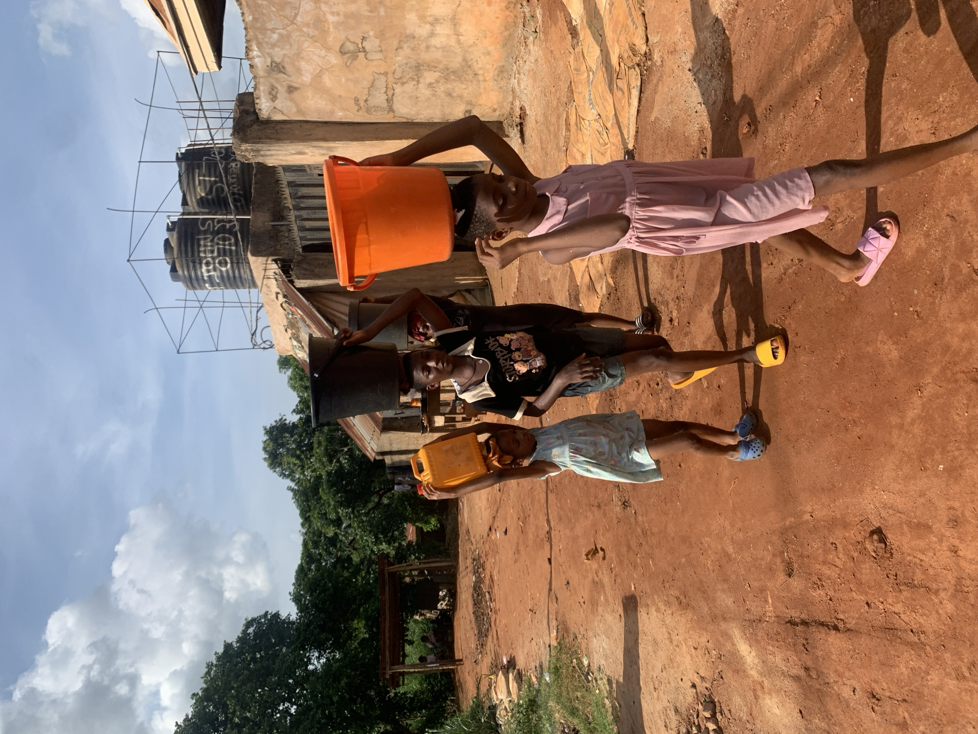 Three children are carrying containers of water on their heads in Ukwu-Abwa, Nigeria. The child on the left is wearing a light blue patterned dress and blue sandals, carrying a yellow water container. The child in the middle is wearing a black graphic t-shirt and gray shorts, with yellow sandals, carrying a black water container. The child on the right is wearing a light pink dress and pink sandals, carrying an orange bucket. In the background, there is a building with black water tanks. The ground is red dirt, and there are trees and a cloudy sky visible.