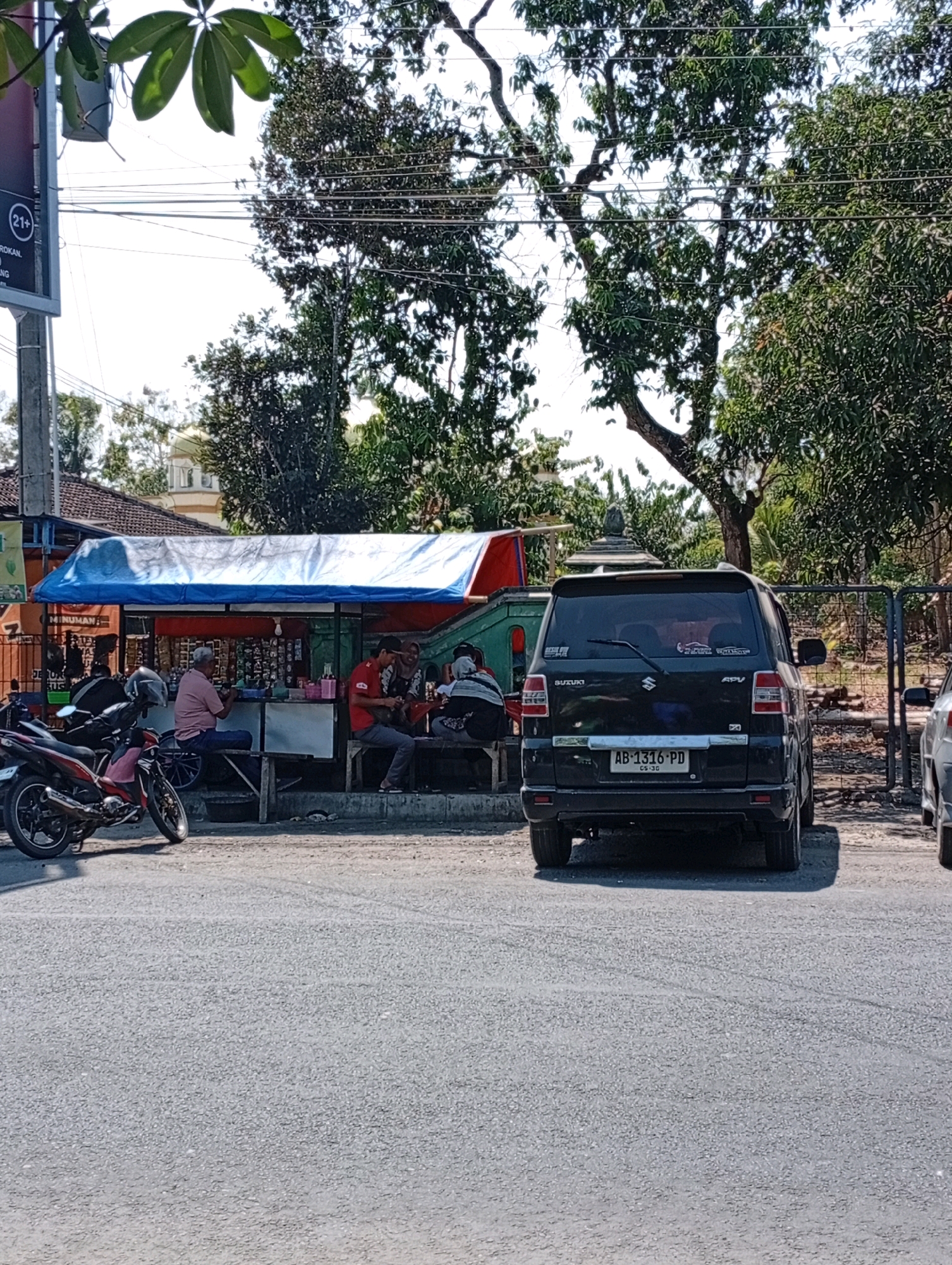 In Playen, Indonesia, a black Suzuki APV van with license plate AB 1316 PD is parked on a paved road in front of a roadside stall. The stall has a blue and red tarp for a roof and displays various items for sale. Three individuals are seated at the stall, two men and one woman, appearing to be interacting with each other. One man, wearing a pink shirt and blue jeans, is seated on a stool facing away from the camera. Another man, in a red polo shirt and dark pants, is seated and engaged in an activity. A woman in a headscarf is also seated with them. To the left of the stall, a red and black motorcycle is parked. Behind the stall, a green utility vehicle is partially visible. Trees and power lines are present in the background, along with a fence. The scene appears to be a typical roadside commercial area.