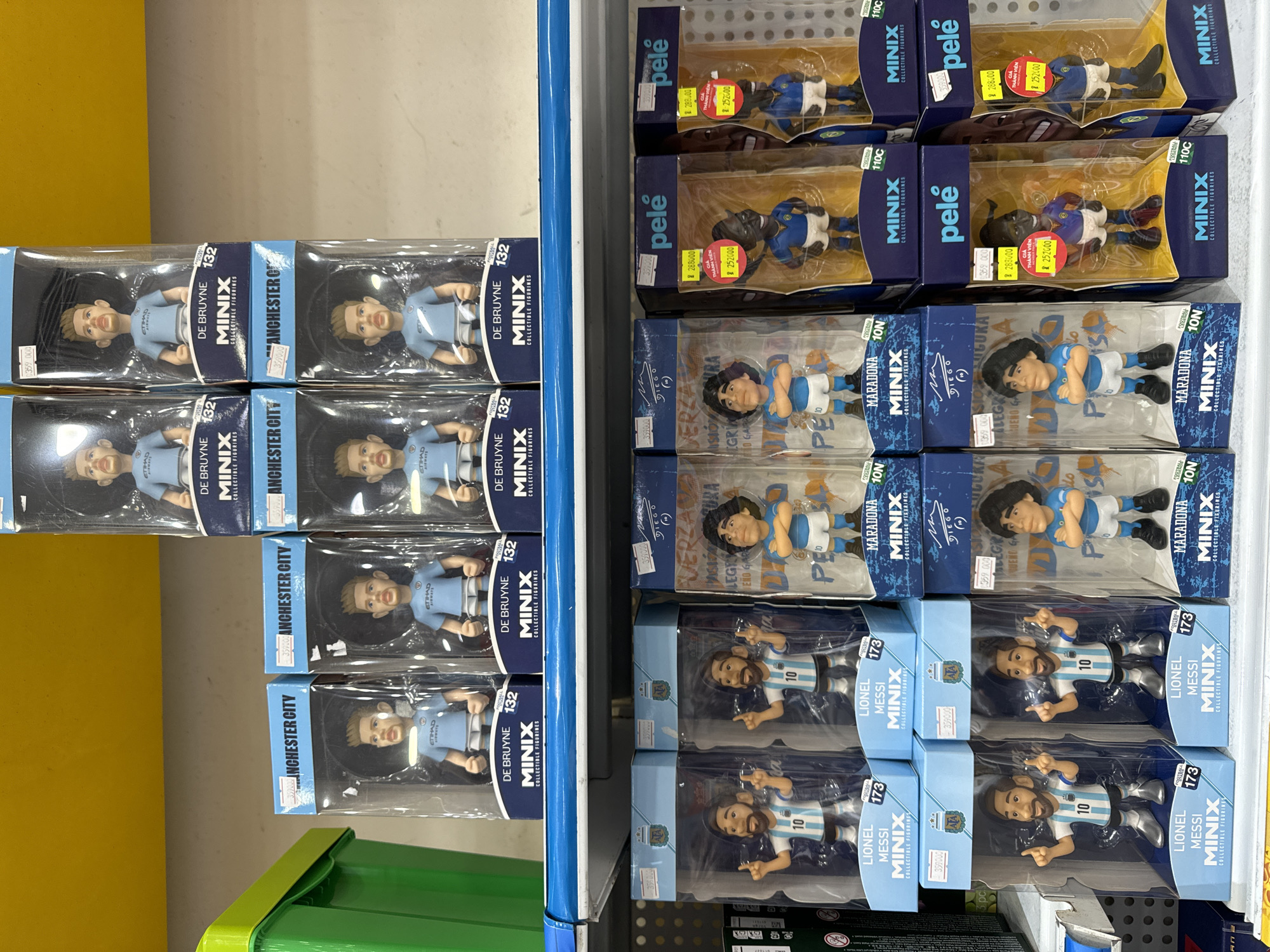 The image shows a retail display in Ho Chi Minh City, Vietnam, featuring Minix collectible figurines of famous soccer players. On the left, there are Kevin De Bruyne figurines in blue Manchester City jerseys. On the right, there are Lionel Messi figurines in blue and white striped Argentina jerseys, Diego Maradona figurines in a light blue jersey, and Pelé figurines. The price tags on each item display prices in Vietnamese Dong. The scene is well-lit, and the shelves appear to be organized.