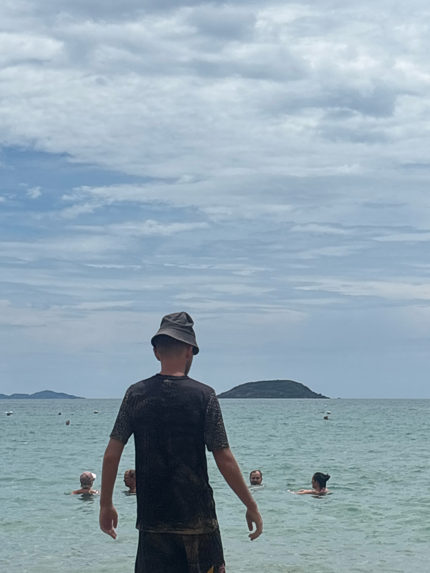 A man wearing a dark bucket hat and a dark t-shirt stands with his back to the camera, facing a body of water. The water is a light turquoise color and appears calm, with gentle ripples. Several people are visible swimming or wading in the water in the mid-ground, including two individuals on the left and two on the right. In the background, a small, dark, tree-covered island is visible in the distance, with a hazy horizon and a sky filled with scattered clouds. This scene is likely located on a beach in Nha Trang, Vietnam, given the typical landscape and the presence of swimmers in the ocean.