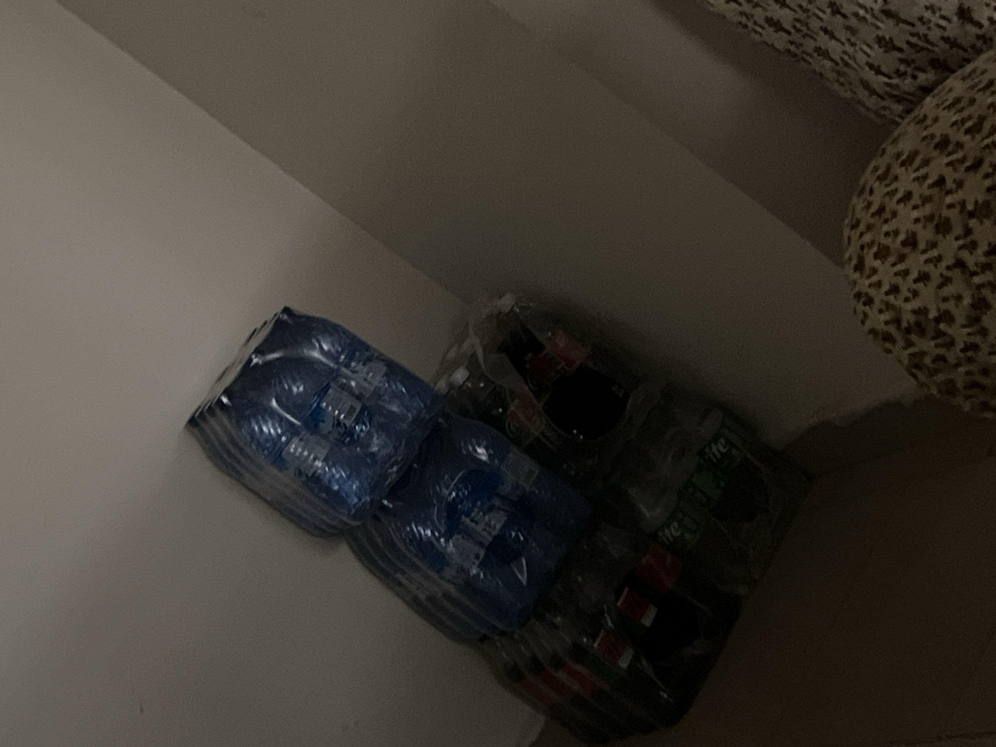 A stack of bottled beverages is leaning against a light-colored wall in a dimly lit room. The bottles are packaged in plastic wrap. The lower portion of the stack appears to contain bottles of Coca-Cola and Sprite, with their labels visible. The upper portion of the stack consists of blue-wrapped bottles, likely also beverages. The overall impression is of a domestic storage area, possibly a pantry or a corner of a room. The provided information states this scene is located in Zawan, Nigeria, though there are no visual cues within the image to confirm this specific location.