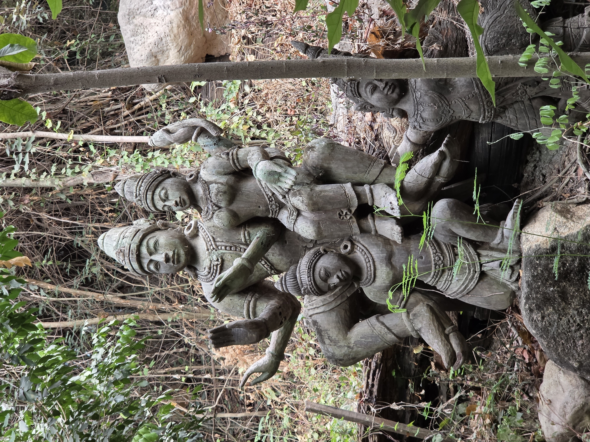 The media file showcases a group of intricately carved statues, likely made of weathered wood or stone, set within a lush, natural environment. There are at least four distinct figures depicted, arranged in a dynamic and interwoven composition. The central grouping consists of three figures: one stands prominently on the left with elaborate headwear and jewelry, its arms gesturing towards a second figure that appears to be reclining or kneeling. A third figure is partially visible below them, contributing to the complex cluster. A fourth figure is seen to the far right, also adorned with traditional headwear, seemingly observing or participating from a slightly lower vantage point.
The activity depicted by the statues is an elaborate, intertwined pose that suggests a narrative, perhaps a dance, a mythological scene, or an embrace. Their expressions are serene. The setting is an outdoor, overgrown area with abundant tropical foliage, including green leaves, dried branches, and visible tree trunks and rocks. The statues exhibit signs of age and exposure, with some areas showing a greenish patina. The lighting suggests it is daytime, with shadows indicating a forested or heavily shaded area. The overall aesthetic is one of ancient artistry nestled within nature. No visible text is present in the image. The scene is located in Pattaya City, Thailand, reflecting a traditional Southeast Asian artistic style.