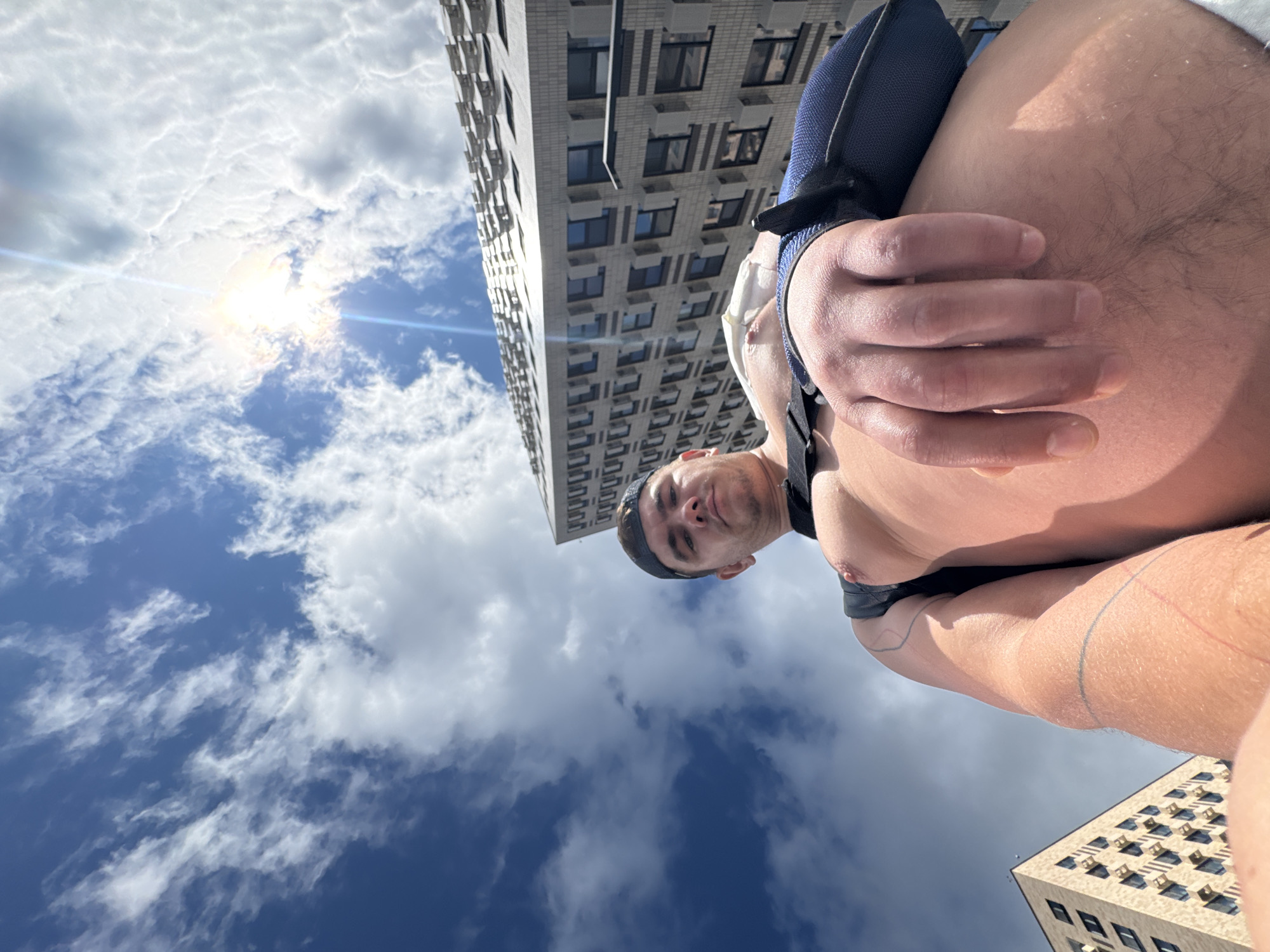 The image shows a low-angle, fisheye perspective of a man, shirtless and wearing a dark blue cap and black shorts, looking up at the camera. He is positioned in the foreground, with his right arm extended and his hand near his face. Behind him, towering apartment buildings with numerous windows are visible against a bright, cloudy sky with the sun glinting. The scene is set in the city of Shcherbinka, Russia, indicated by the distinctive architecture of the buildings, which are characteristic of residential complexes in the Moscow Oblast. The man appears to be in an outdoor urban environment, possibly a courtyard or street area between buildings.