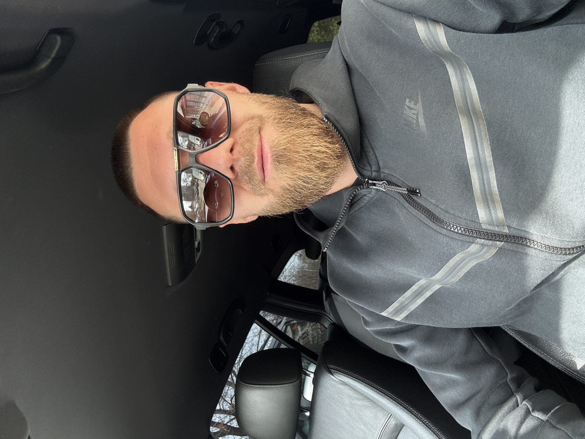 A fair-skinned male with short brown hair and a light beard, wearing sunglasses and a grey Nike hooded sweatshirt with silver stripes, sits in the driver's seat of a vehicle with black leather seats and a black interior. The reflection in the sunglasses suggests an outdoor urban setting. Based on the architecture and vegetation visible in the reflection, the location is potentially in Sofia, Bulgaria.