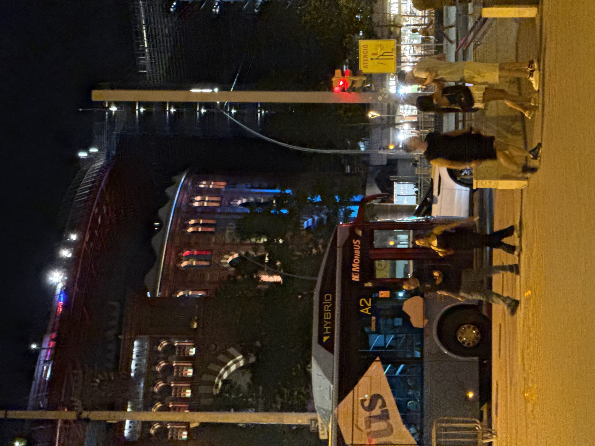 In this nighttime scene in Barcelona, Spain, likely near the Olympic Port area, a "MONBUS" hybrid bus is parked on a street. The bus is gray with yellow and black accents, displaying "US" on its side and "A2" as a route number. To the right of the bus, a traffic light is red. A yellow sign with black text reading "ATENCIÓ" (attention in Catalan) is also visible.
Three individuals are walking on the pavement. The first, a male in a black sleeveless shirt and gray pants, is walking away from the camera. The second, a female in a dark dress, is walking alongside him. The third, a male wearing a light-colored t-shirt and light shorts, is walking in the same direction, interacting with another person not fully visible in the frame.
In the background, a large, multi-story building with arched windows is illuminated, and a structure resembling a bridge or elevated walkway with red and blue lights spans overhead. The overall atmosphere is that of a bustling urban environment at night.