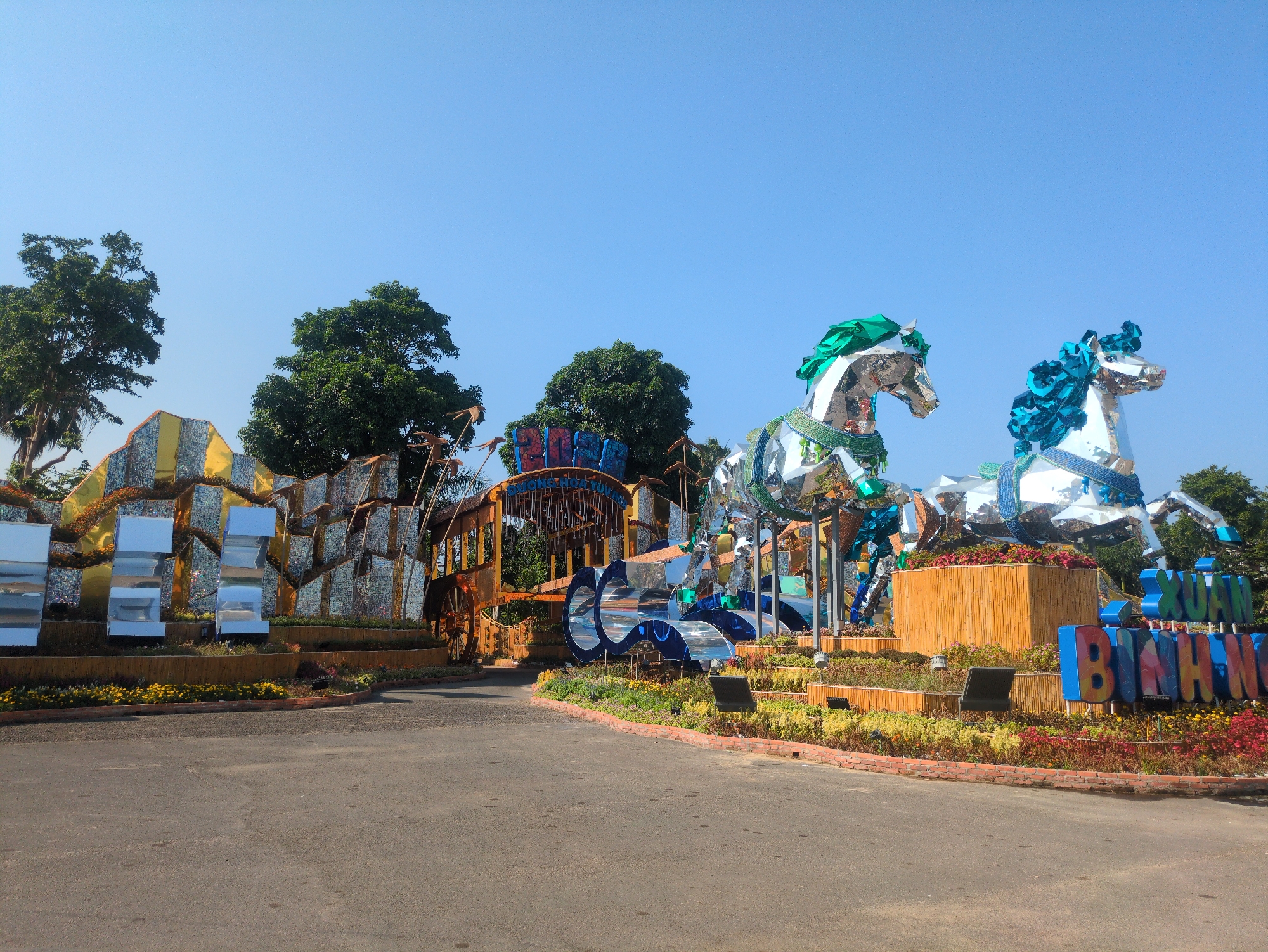 The image captures a street scene in Tuy Hòa, Vietnam, featuring elaborate floral arrangements and sculptural installations. Dominating the foreground are two mirrored horse sculptures with green and blue accents, respectively, set atop raised platforms. Behind them stands a structure resembling a carriage, adorned with more mirrored elements and positioned to appear as if passing through an archway marked with the name "XUAN BINH HOA" in colorful, oversized letters. The street is paved and curves gently towards the entrance of the display. The sky is clear and blue, with trees scattered in the background. The overall impression is of a festive, decorative display, likely part of a local celebration or event.