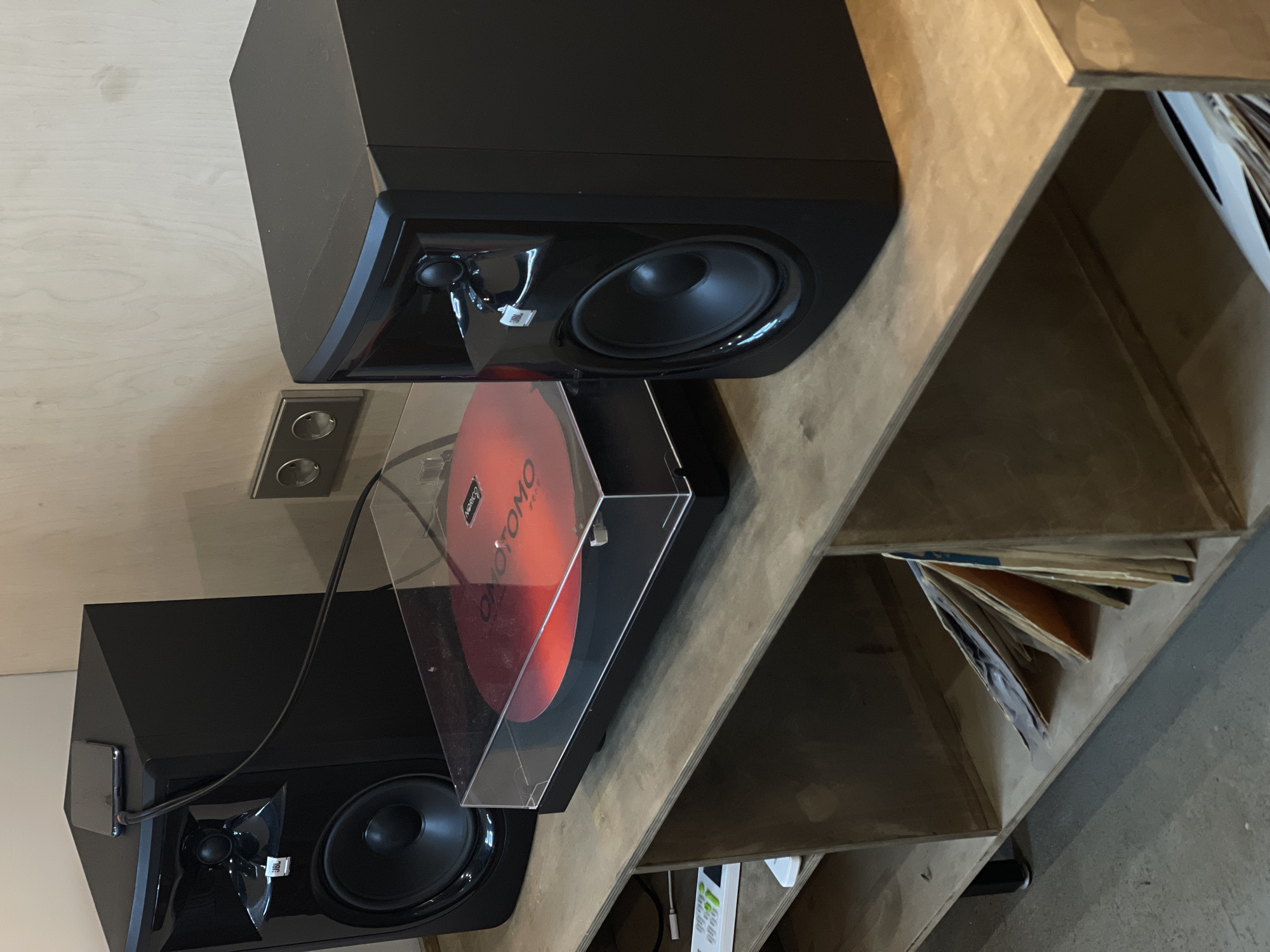 An indoor, eye-level, medium shot shows a turntable and two black speakers, positioned on a wooden shelf unit. The turntable is centered on the shelf, with a red record playing. A smartphone is placed on top of the speaker to the left. To the right, another speaker is positioned with a wall outlet visible behind it. The shelf unit contains several records stored vertically on the lower shelf. The wall behind the unit is a light-colored wood paneling. The lighting is subdued and natural.