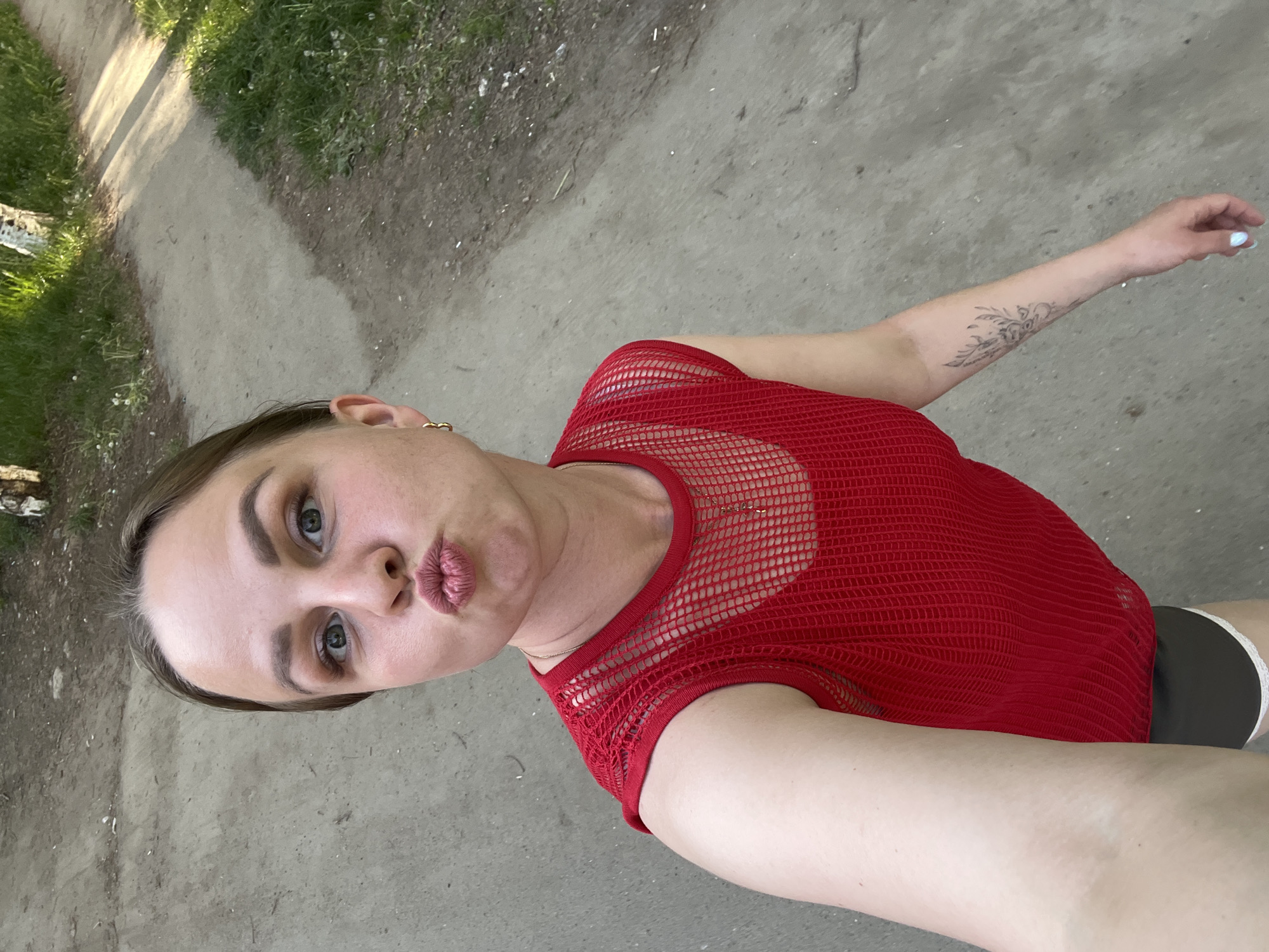 A woman stands outside on a paved path in daylight. She is wearing a red mesh top, gray athletic shorts, and has a tattoo on her left forearm. She is making a kissing face towards the camera, which is tilted at an angle. The path is bordered by grass and some trees. The location is possibly in Izhevsk, Russia, based on the prompt's specification.