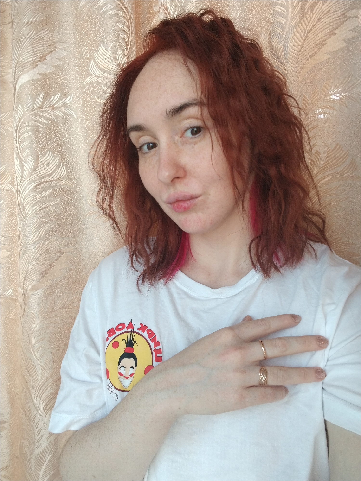 A young, light-skinned woman with red, wavy hair poses for a selfie indoors, likely in Izhevsk, Russia, due to the design on her t-shirt. The t-shirt features a cartoon image inside a yellow circle with the text "ТИНК WINK OBOK" in red around the circle. The woman has a ring on each ring finger and her hand rests on her chest. A patterned curtain is in the background.