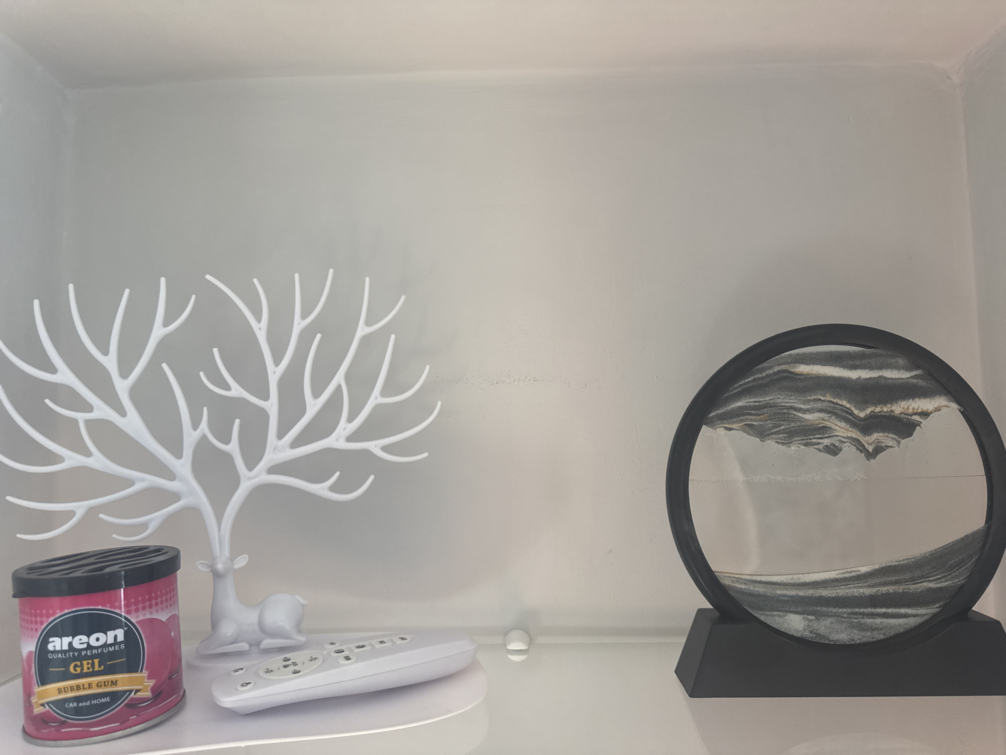 The image shows a shelf in a white interior, possibly a built-in cabinet. From left to right, the shelf contains: an "areon QUALITY PERFUMES GEL BUBBLE GUM CAR and HOME" air freshener, a white deer-shaped jewelry stand, a white remote control, and a black sand art display on a base. There are no visible people or indications of a specific location in Kryvyi Rih. The focus is on the arrangement of objects on the shelf.