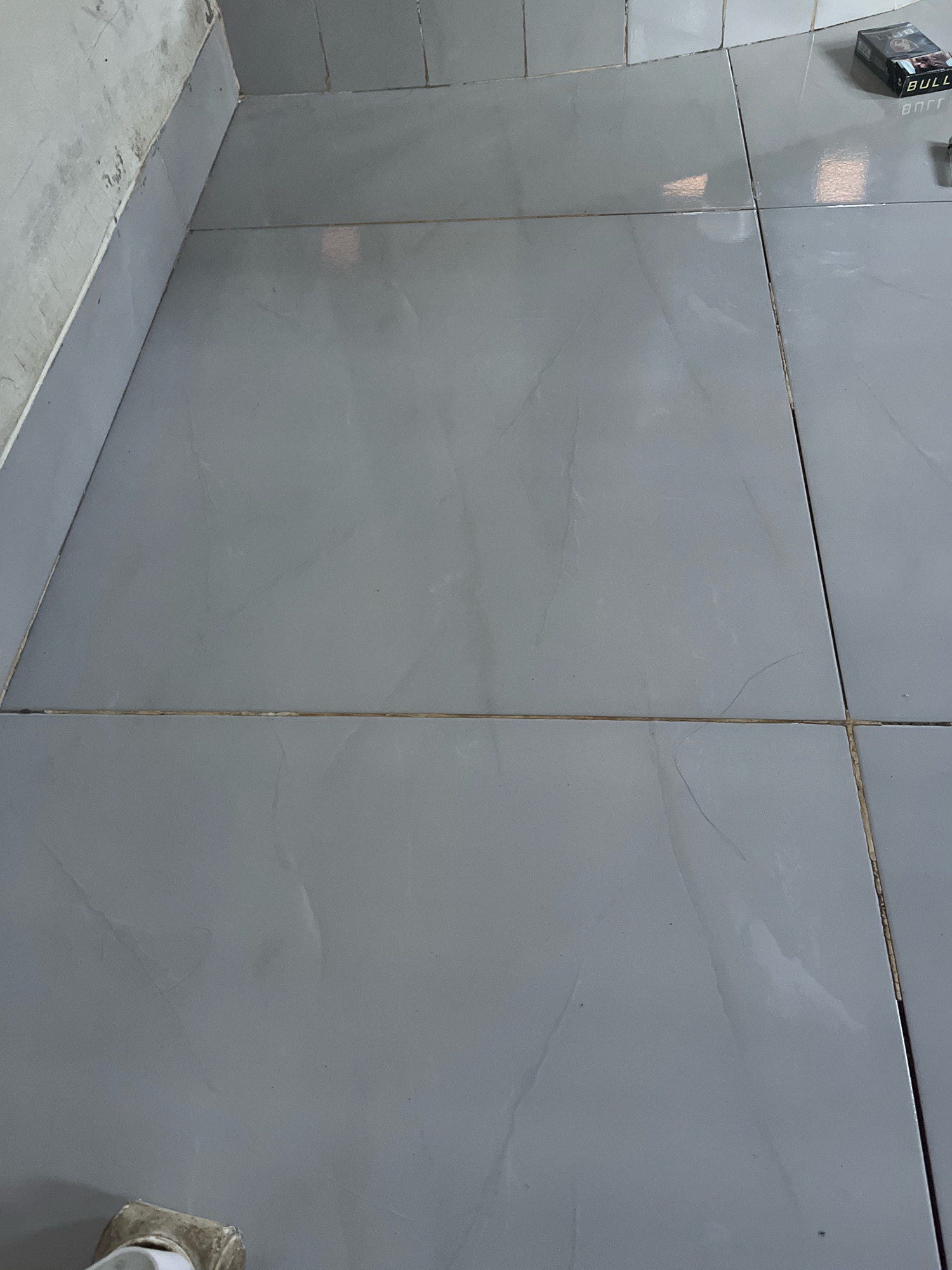 A pack of "BULL" brand cigarettes is placed on a tiled floor. The tiles are large, square, and light gray with faint veining. The grout lines are visible and appear to be a light brown color. The scene is lit from above, casting reflections on the glossy surface of the tiles. The environment appears to be indoors, possibly a room or hallway. There is no indication of the specific location in Kebumen, Indonesia, beyond the general appearance of the floor and the presence of the cigarette pack.