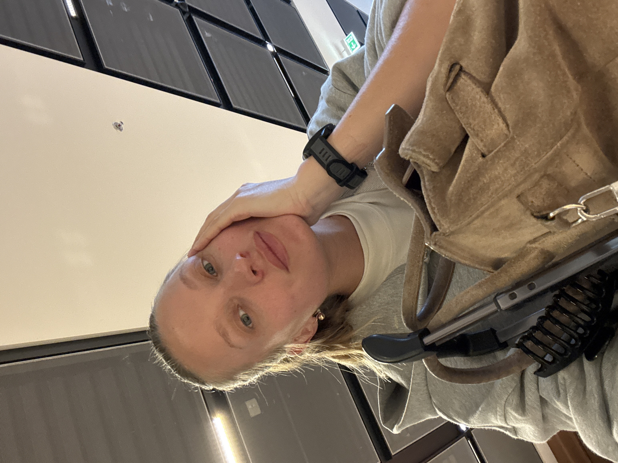 In Schönefeld, Germany, a woman with blonde hair pulled back in a ponytail and minimal makeup is taking a selfie. She is wearing a gray hooded sweatshirt over a white t-shirt and a black digital watch on her left wrist. Her right hand is resting on her cheek. In the foreground, a light brown suede bag with silver hardware is visible, along with a black stroller handle. The background appears to be an indoor space with a ceiling featuring geometric panels and fluorescent lighting. A green exit sign is visible on the right side of the frame.