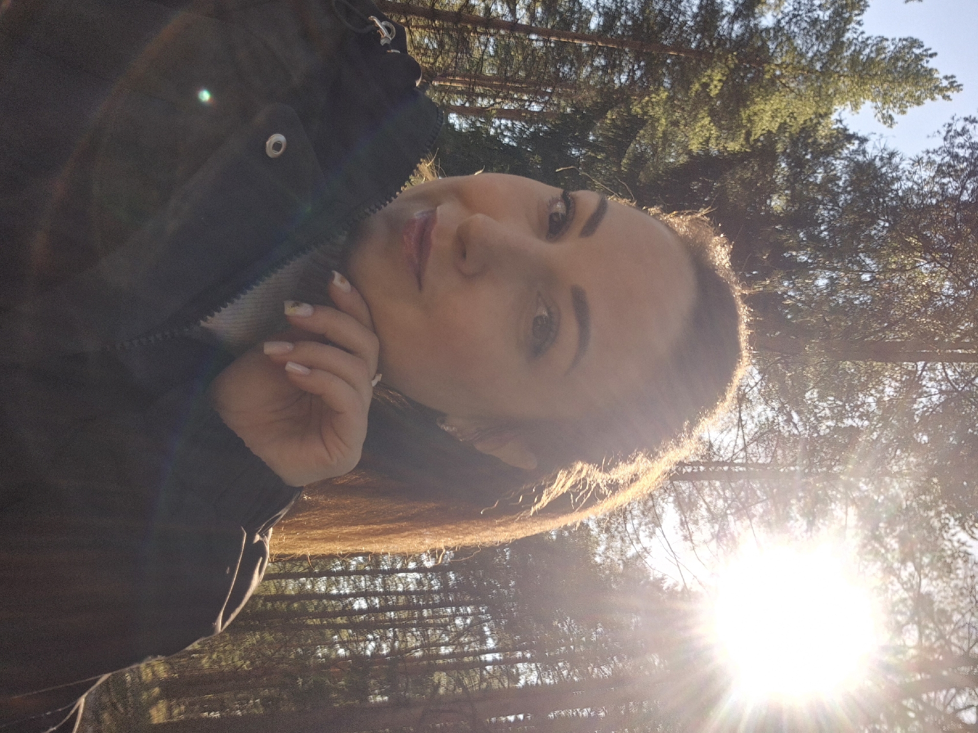 A portrait-oriented selfie captures a young woman outdoors in a sunlit forest. The woman, positioned on the left side of the frame, has long, dark hair, fair skin, and is looking directly at the camera with a gentle expression. Her hair is heavily backlit by the sun, creating a bright halo effect around her head. She is wearing a dark, possibly padded jacket or coat with a visible zipper and a silver-colored snap button on the lapel. Her right hand is gently placed near her chin/cheek, showcasing manicured nails with a light polish.
The background consists of numerous tall pine trees, their dark green foliage contrasting with the bright sky. A strong, radiant sunburst dominates the lower right portion of the image, causing significant lens flare and dappled light throughout the scene. The clear, bright sky is visible above the trees. The lighting suggests either early morning or late afternoon, often referred to as "golden hour," given the low angle of the sun and the warm, intense glow it casts. The overall mood is serene and natural. There is no visible text in the image.