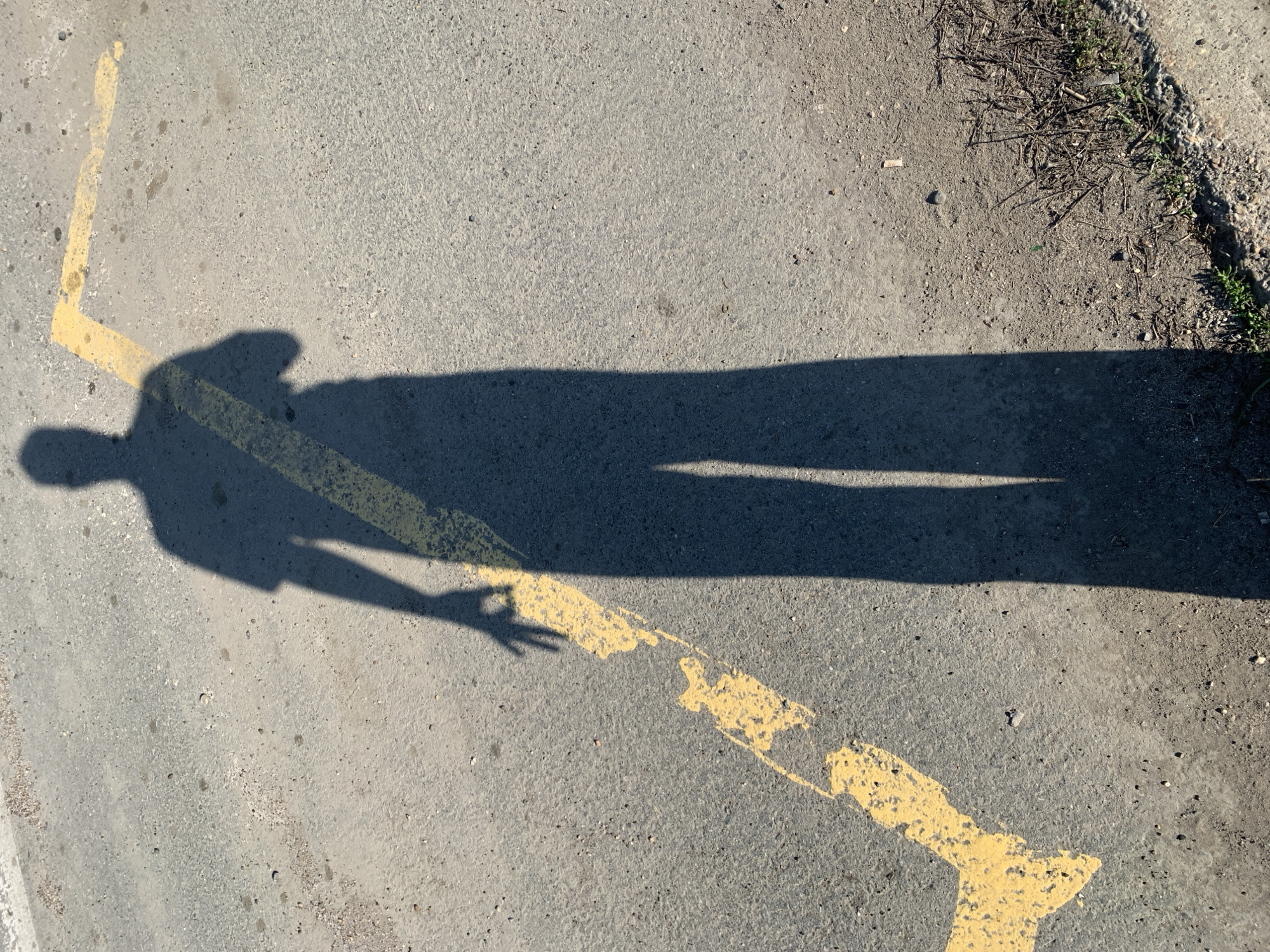 The image shows a shadow of a person on a gray paved surface. The shadow is elongated and stretches across a fragmented yellow painted line on the ground. The shadow appears to be gesturing with one hand, holding up two fingers. To the upper right, there is an edge of unpaved ground with small plants and debris. The specific location in Chaikovsky, Russia cannot be determined solely from this image.