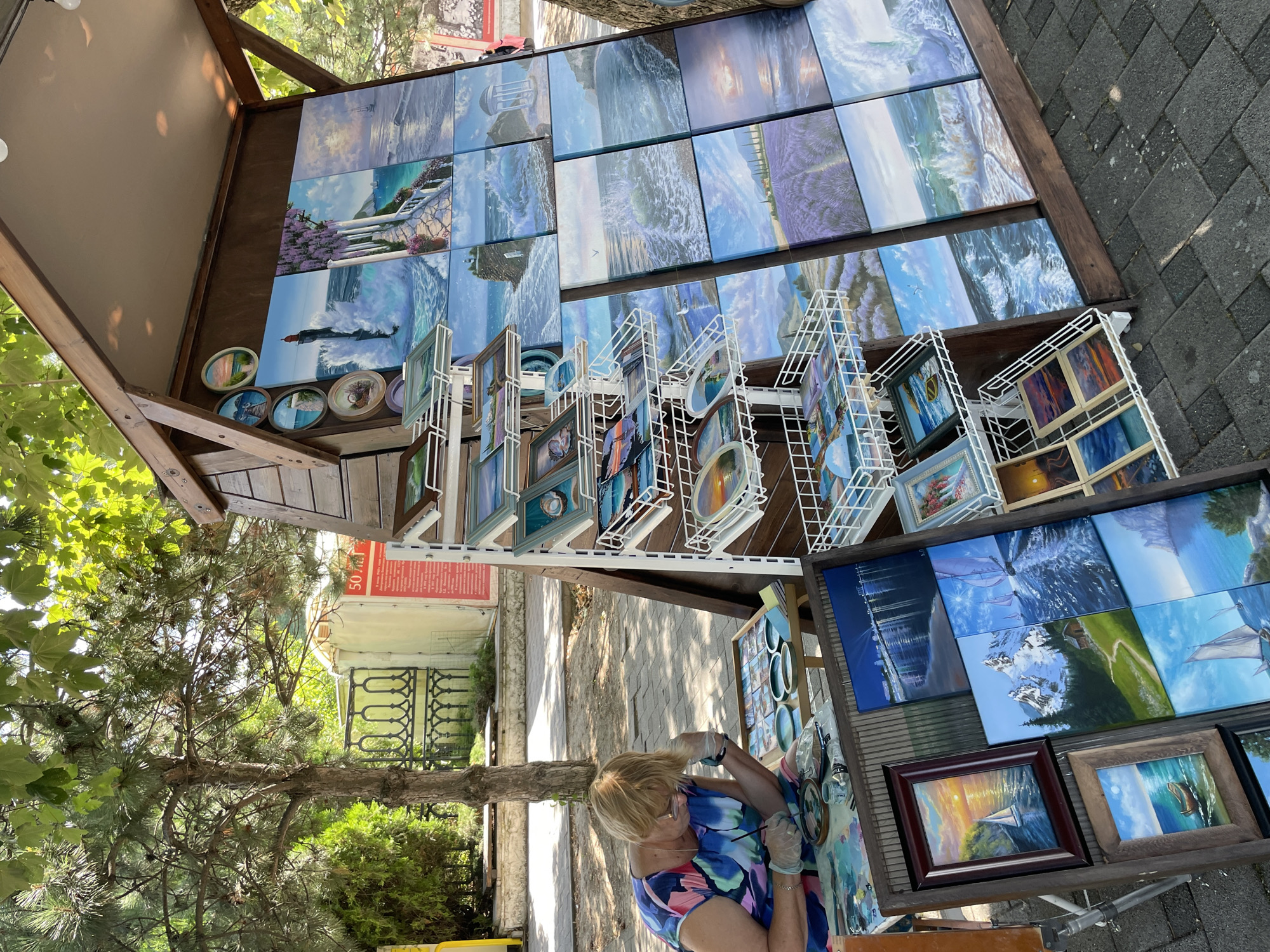 In Anapa, Russia, an artist is painting outdoors. She is seated at a table, wearing a colorful dress and gloves, and appears to be working on small circular paintings. In front of her, various art supplies are spread out, including paints, brushes, and small canvases. Behind her is a wooden display structure showcasing a collection of landscape paintings, primarily featuring seascapes, lavender fields, and coastal scenes. The artwork is arranged on shelves and leaning against the structure. A tree with green foliage provides shade over the scene, and the ground is paved with grey stones. In the background, a fence and a wall with a red sign are visible.