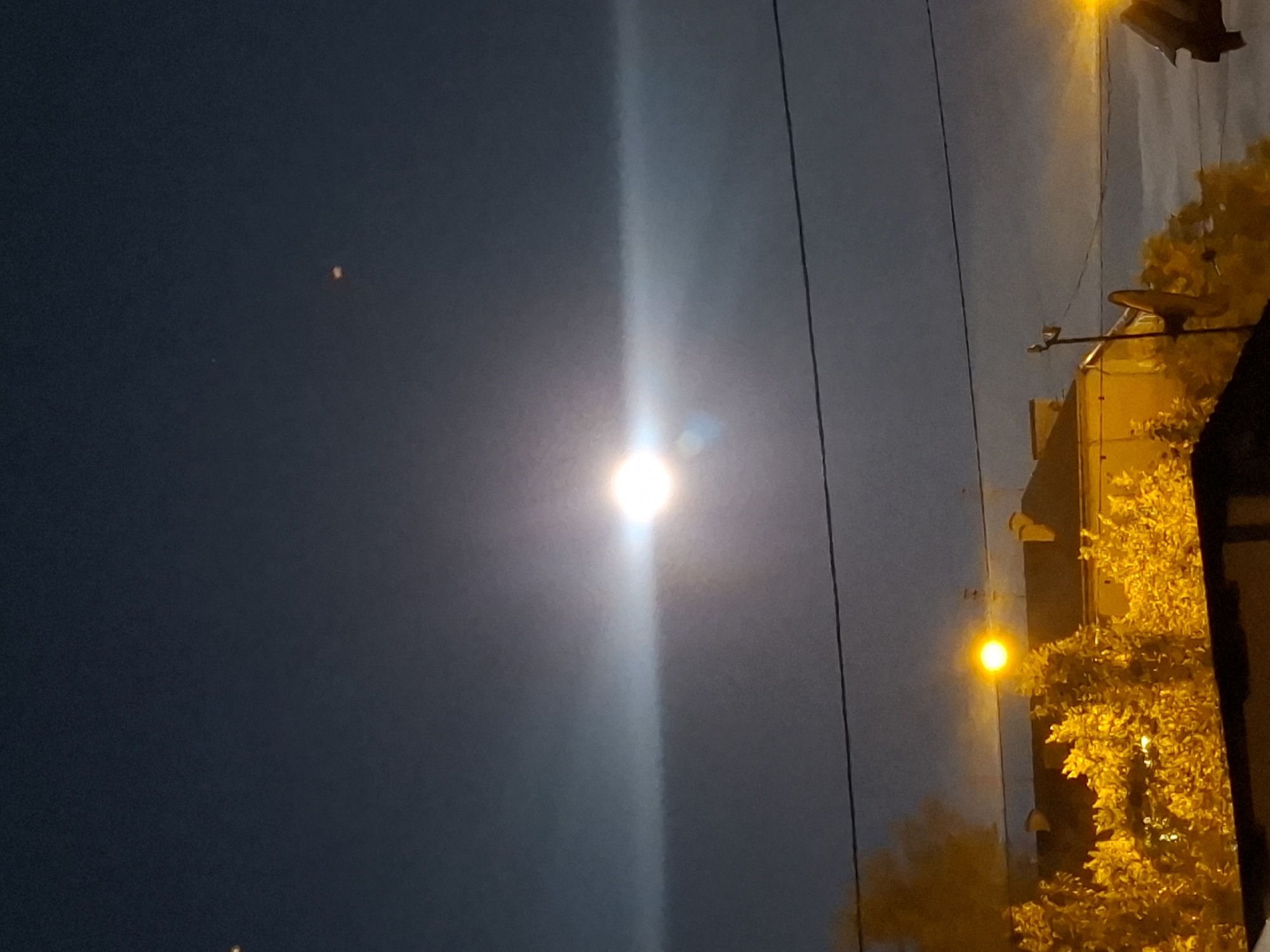 The image shows a night sky over the city of Tyumen, Russia, with a bright light source in the center, possibly the moon or a street lamp, creating a lens flare effect that extends horizontally across the frame. There are also two smaller, indistinct points of light higher in the sky. In the lower portion of the image, rooftops of buildings are visible, illuminated by artificial lights, along with trees or foliage also lit up with a warm, yellow glow. Several power lines stretch horizontally across the scene. The overall impression is of a nocturnal urban landscape.