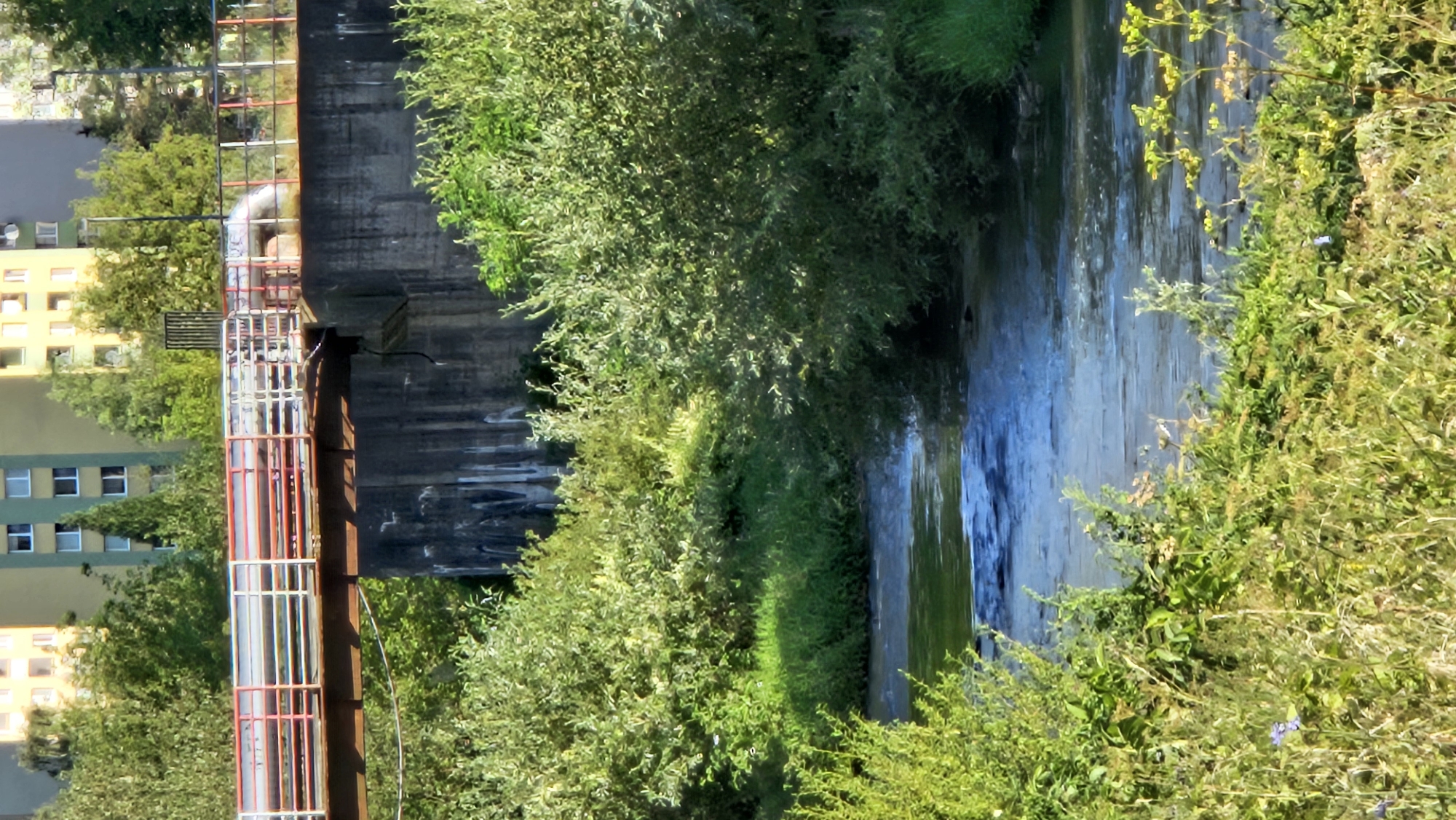 The image depicts a river flowing through a green, overgrown area with a concrete bridge overhead. The bridge supports a large pipe, suggesting an industrial or infrastructure element within the urban landscape. In the background, multi-story residential buildings are visible, characteristic of a city. The scene is set in Prešov, Slovakia, a city known for its architectural diversity and proximity to natural elements within its urban planning.