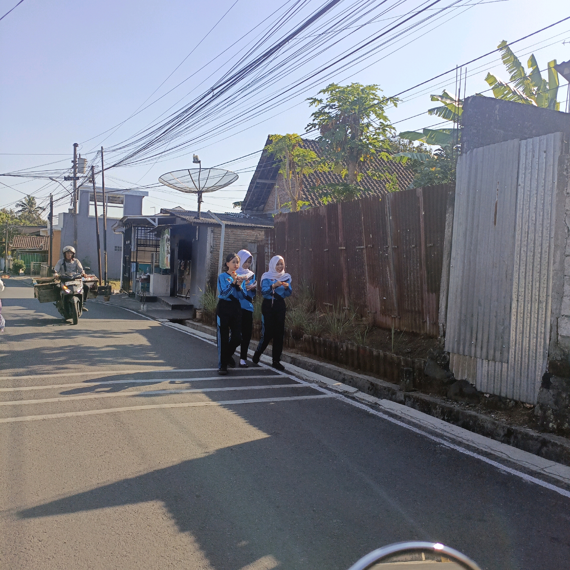 In Salatiga, Indonesia, on a sunlit day, three young women in blue and black uniforms, wearing hijabs, walk across a paved road marked with white pedestrian stripes. They appear to be holding small objects. To their left, a man on a motorbike, wearing a helmet and a grey shirt, rides past carrying a wooden crate attached to the front. Behind the women, a corrugated metal fence runs along the side of the road, bordered by vegetation and a small ditch. The background shows a residential area with buildings, power lines crisscrossing overhead, and a satellite dish on a pole. The road itself is asphalt, with shadows cast across it from the surrounding environment.