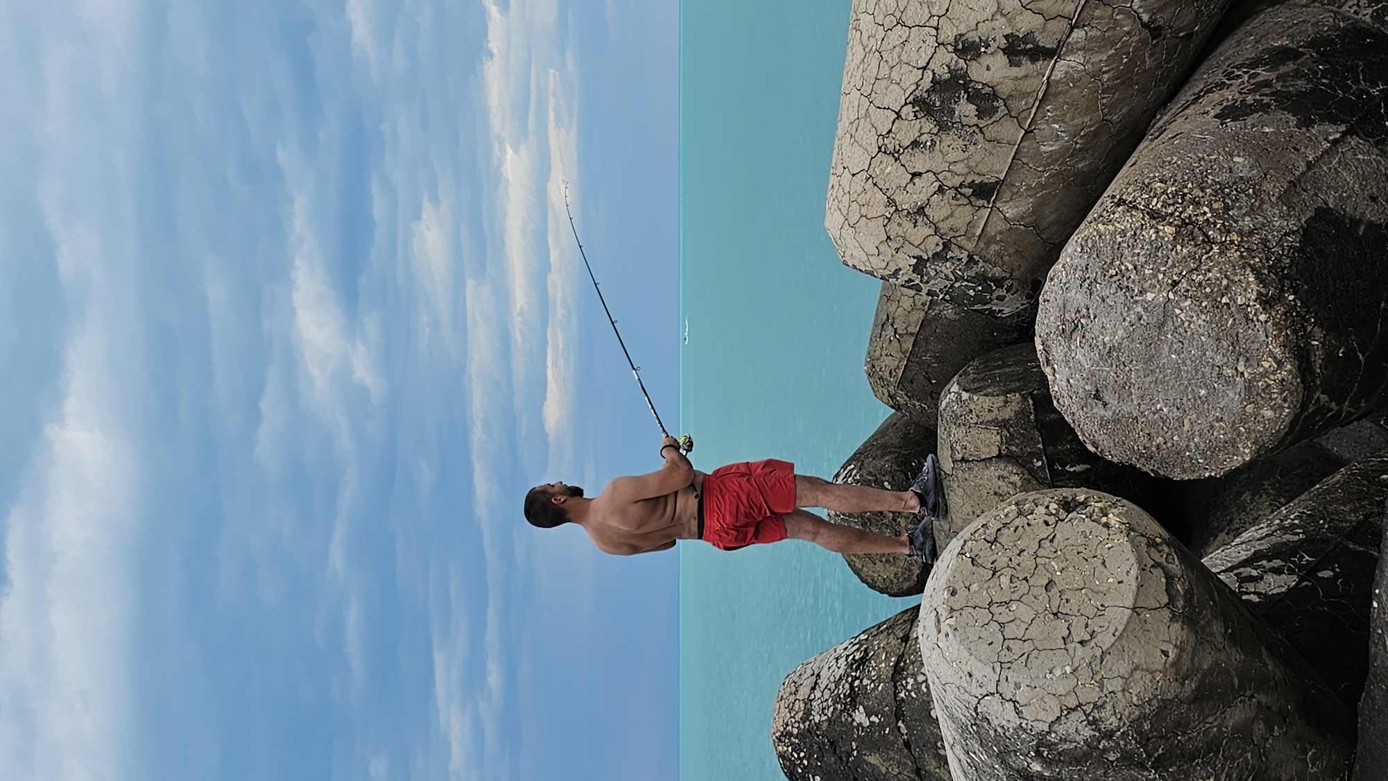 A man is fishing from a breakwater on the coast. The man is fair-skinned with dark hair and a beard, wearing red shorts and dark-colored sneakers. He is holding a fishing rod, which extends toward the turquoise sea. The sky is light blue with wispy clouds. The breakwater is composed of large, cylindrical concrete structures with cracks and discoloration. The location is likely Varna, Bulgaria due to the prevalence of such breakwaters along the Black Sea coast.