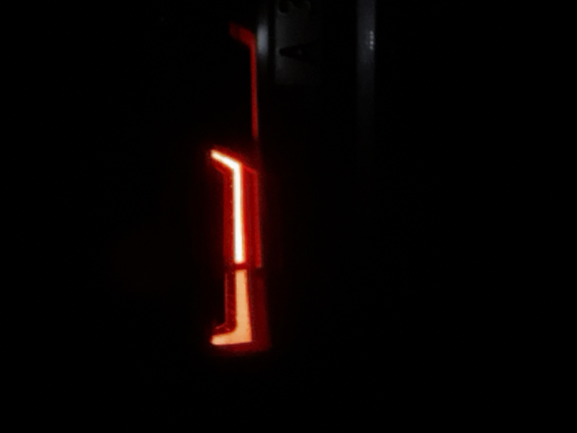 The image displays a dark, low-light environment featuring the illuminated rear section of a vehicle. A prominent red-orange LED taillight assembly is visible, characterized by a distinct L-shaped element on the left transitioning into a horizontal light bar extending rightward. Above this main light bar, a thinner, shorter light strip is present on the right side. Below and to the right of the primary light assembly, a rectangular vehicle license plate is partially visible. The characters "A 3" are faintly discernible on the illuminated portion of the plate. No individuals or other distinct objects are visible in the uniformly dark background. The visible elements are consistent with the rear exterior of a passenger vehicle. This scene is located in the city of Varna, Bulgaria, indicated by the "A" regional code on the license plate.