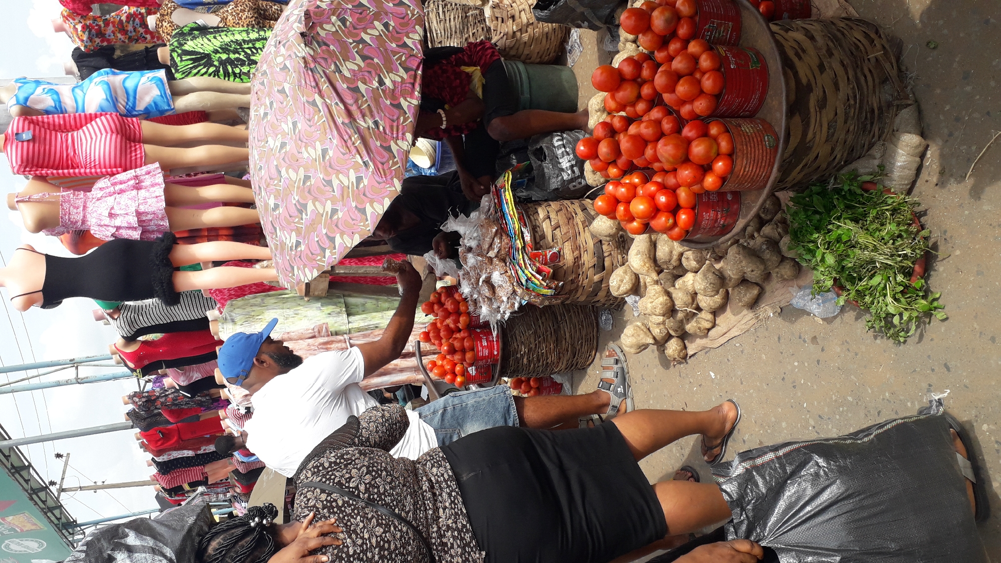 The image captures a vibrant scene at the Onitsha Main Market in Onitsha, Nigeria, a bustling hub of commerce. In the foreground, a woman dressed in a black skirt and a patterned blouse stands on the left, holding a large black plastic bag. Beside her, a man wearing a blue cap and a white t-shirt is inspecting produce. Piles of yams and tomatoes are displayed for sale, arranged in baskets and on a metal stand. To the right, two women are engaged in a transaction under a colorful umbrella. The background reveals rows of mannequins adorned with various colorful dresses, typical of clothing stalls in the market. The ground is paved, and the overall atmosphere suggests a busy market day with a focus on fresh produce and clothing.