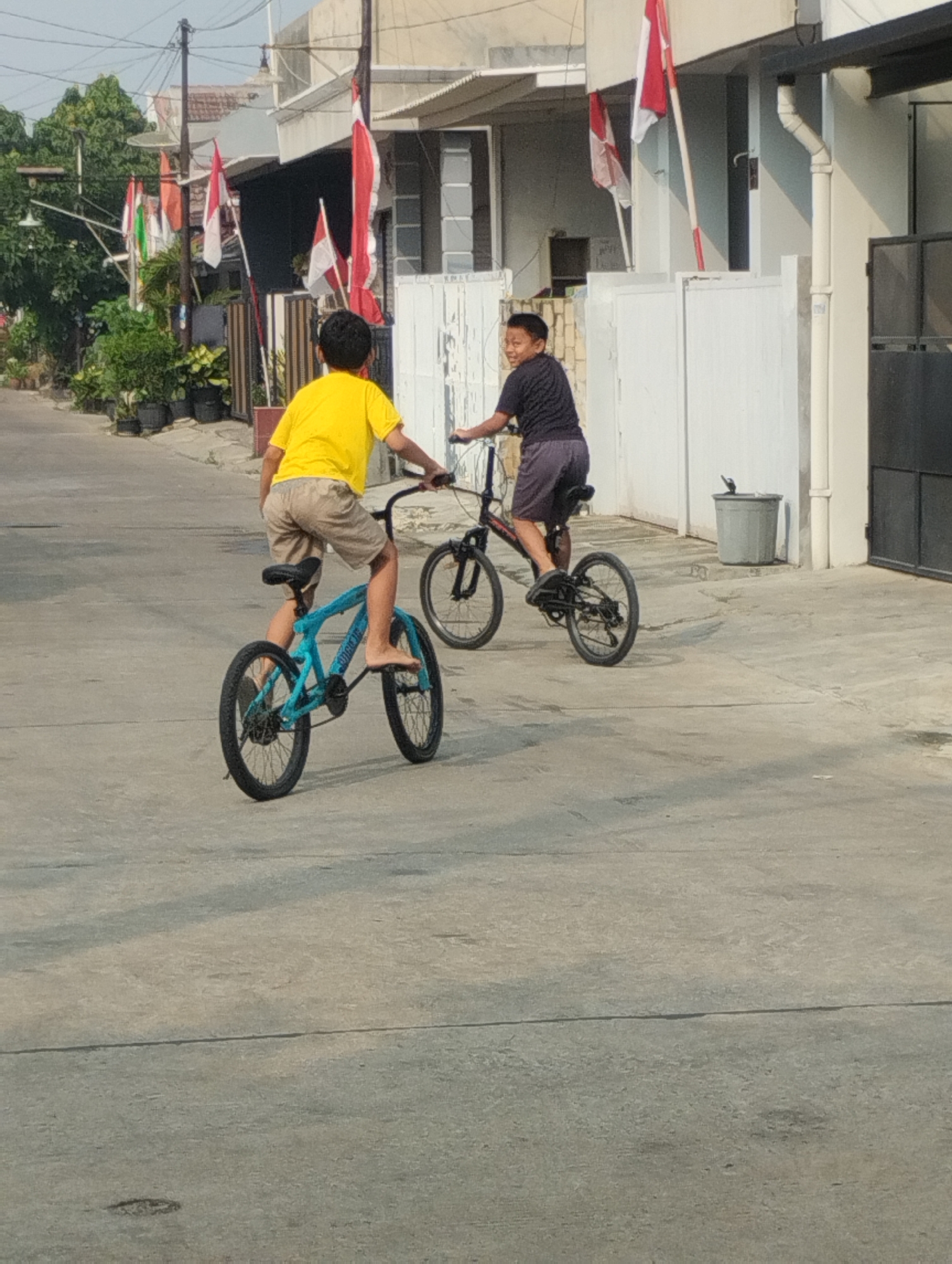 Two young boys are riding bicycles on a paved street in front of residential buildings in Bekasi, Indonesia. The boy in the foreground is wearing a yellow t-shirt and light-colored shorts and is riding a blue bicycle. The boy in the background is wearing a dark t-shirt and dark shorts and is riding a black bicycle. Indonesian flags are visible in the background, suggesting a national holiday or event. The environment is sunny with some trees and potted plants visible.