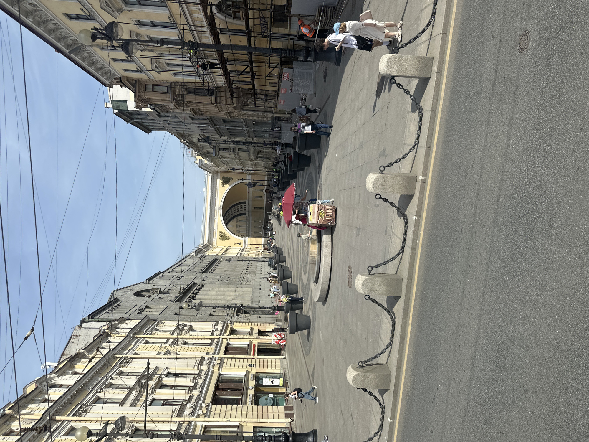 This image shows a street in Saint Petersburg, Russia. The view is an axial perspective of Palace Square facing the General Staff Building with the Triumphal Arch. Buildings line each side of the square, where people are walking. On the right side of the frame, the street borders the road. A row of concrete blocks connected by chains runs along the edge of the sidewalk. Further down the street, a merchant is selling goods from under a large, red umbrella. The overall impression of the scene is a bright, sunny day.