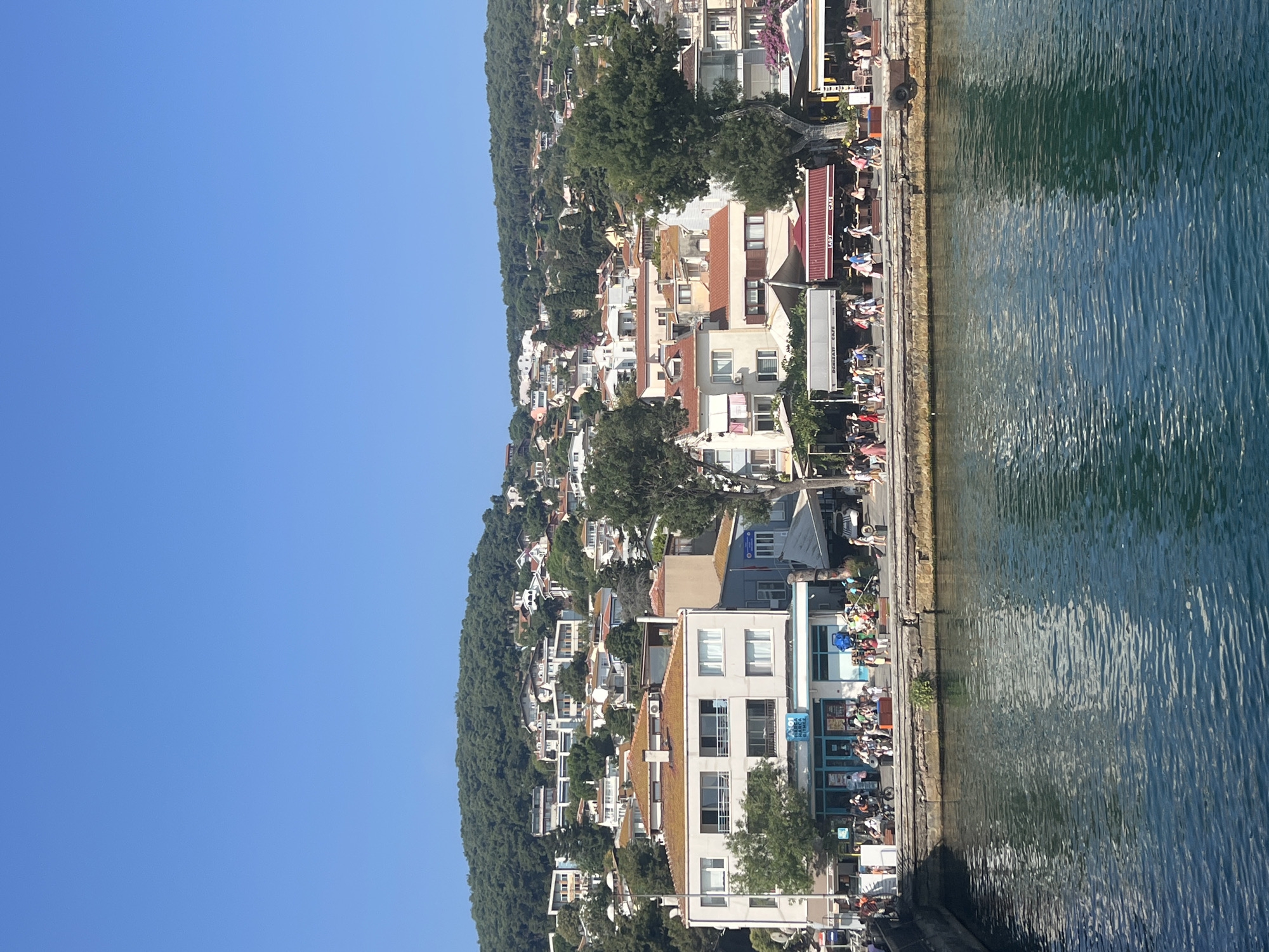A bright, sunny day is depicted with a clear blue sky. In the foreground, a body of deep blue water with gentle ripples fills the lower portion of the frame. Along the edge of the water, a stone embankment is visible. Above the embankment, a pedestrian promenade runs alongside a row of buildings. These buildings are primarily two to three stories high, with many featuring reddish-brown tile roofs. Some buildings have awnings and outdoor seating areas, suggesting they are cafes or shops. A diverse group of people, some walking individually and others in small groups, are strolling along the promenade. They are dressed in casual summer attire. Further inland, the scene transitions to a hillside densely covered with green trees and vegetation. Scattered amongst the trees are more buildings, also with reddish-brown roofs, climbing up the slope. The architecture suggests a coastal town or island setting. One building near the front, with white walls and multiple windows, appears to be a commercial establishment with signage. The overall impression is a lively waterfront scene on a warm day in a Mediterranean or Aegean coastal town.