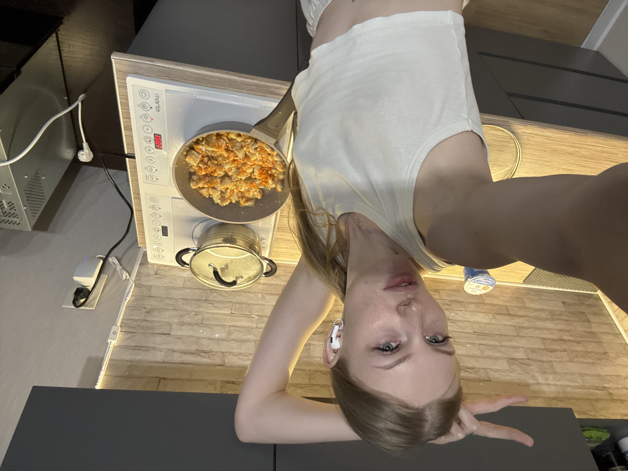 The image is an upside-down selfie of a woman with long blonde hair, wearing a white tank top and a wireless earbud in her left ear. She is making a peace sign with her right hand. In the background is a kitchen environment. On the stovetop, there is a frying pan with food that appears to be scrambled eggs with some red pepper flakes and a pot with a lid. The stove is a "Marta" brand induction cooktop. A brick-patterned backsplash is visible. Other visible objects include a computer case and a kitchen counter. The location is the city of Perm, Russia.