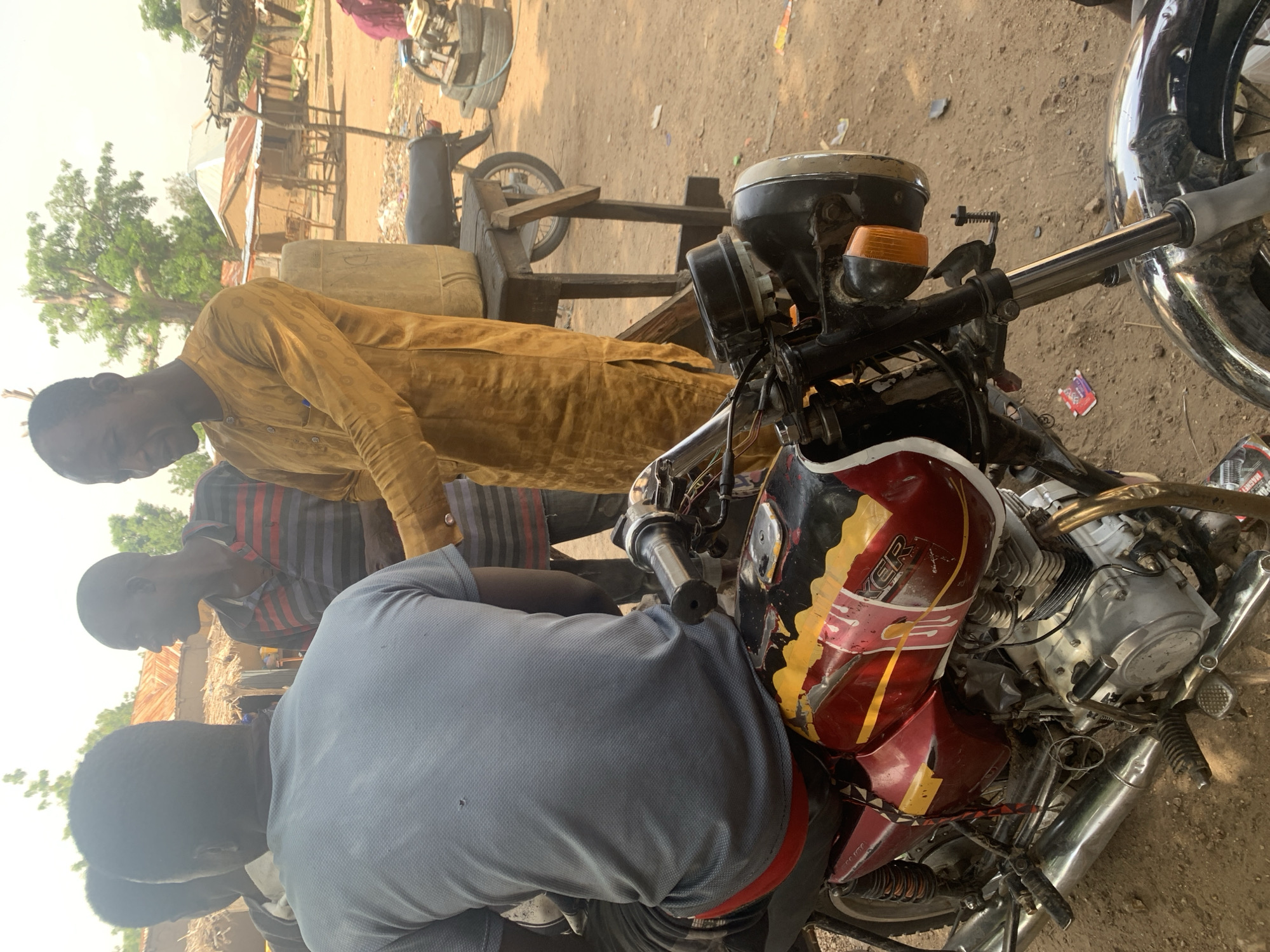 In Gobbiya, Nigeria, a group of men are gathered outdoors near a dirt road, with several buildings and trees visible in the background. The focus of the image is a motorcycle parked in the foreground. A man wearing a yellow traditional outfit is standing next to the motorcycle, appearing to interact with another man who is seated on the motorcycle, facing away from the camera and wearing a grey t-shirt and red shorts. Another man in a striped shirt is also present, looking towards the interaction. In the background, a rough wooden table holds a large plastic container. The scene depicts a casual gathering in what appears to be a rural or semi-urban setting.