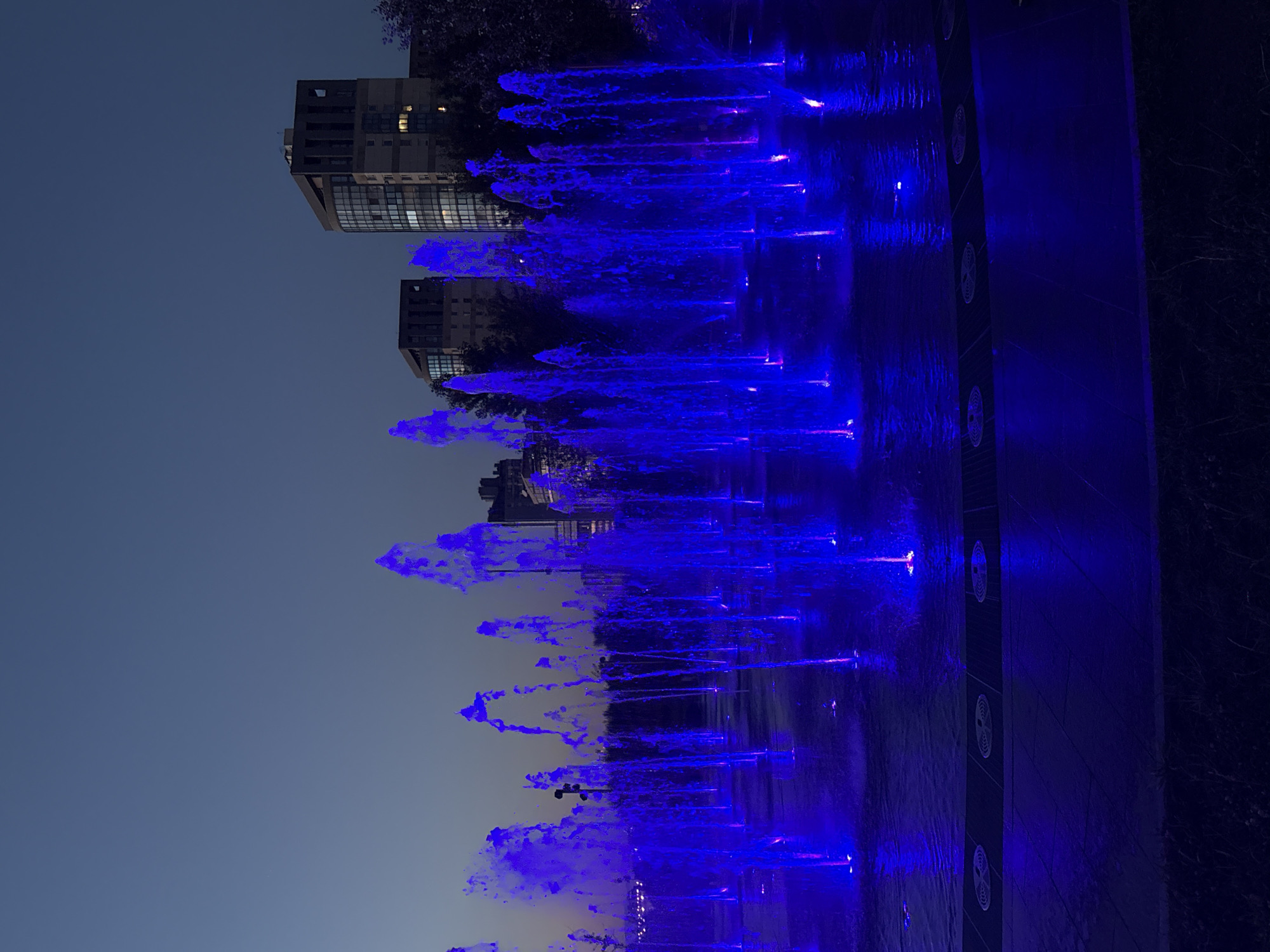 This is a nighttime photograph of a water fountain display in Saint Petersburg, Russia. The water jets are illuminated with vibrant blue lights, creating a dynamic and visually striking effect. The fountain is located in a paved area, and in the background, several modern buildings are visible, their windows illuminated. The sky is a deep twilight blue. There are no people visible in the image.