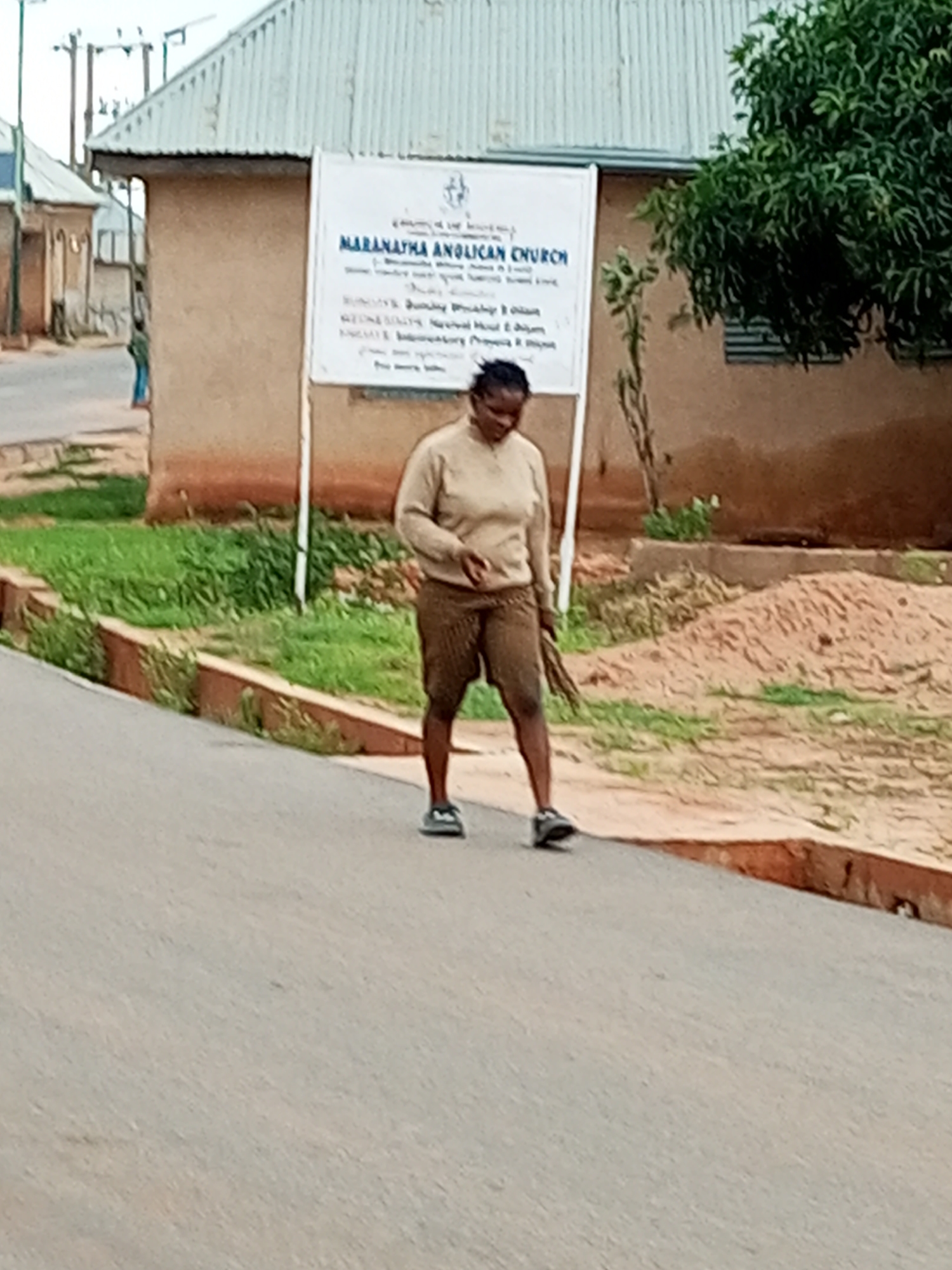 An adult female is walking on a paved road in Gombe, Nigeria. She is wearing a tan sweater and patterned shorts. She is holding a bundle of sticks or branches in her right hand. To the left of the woman, there is a building with a sign that reads "MARANATHA ANGLICAN CHURCH." The building appears to be made of earth or stucco and has a corrugated metal roof. In the background, there is green grass and a large tree with lush foliage. The road is lined with a low brick border. The sky is overcast.