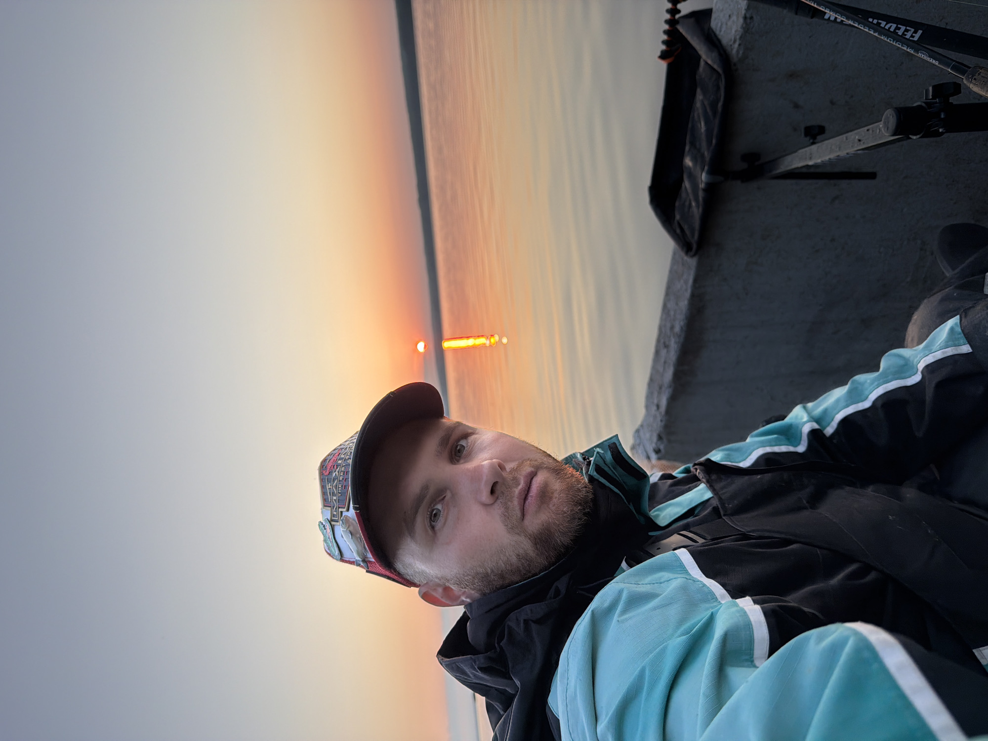 A man with a beard and a baseball cap is taking a selfie by a body of water during sunset. He is wearing a teal and black jacket. In the background, the sun is setting over the water, casting an orange glow. A concrete barrier and fishing rods are visible in the foreground. The image appears to have been taken in Chaikovsky, Russia, given the context provided, likely near the Kama River.