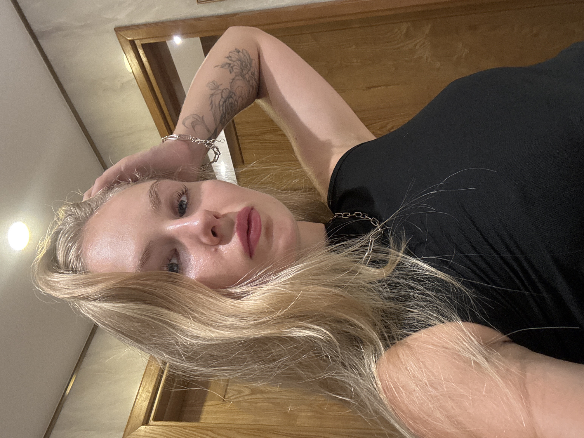 A young blonde woman is captured in a close-up, selfie-style shot. She is indoors, with her left hand casually resting on her head, and her right arm bent upwards, exposing her left arm which has a tattoo. The woman is wearing a black tank top and a silver chain necklace. Her gaze is directed at the camera, and her lips are slightly parted. The background appears to be a hallway with wooden doors, suggesting a residential or hotel setting. There are no explicit indicators to place the scene in Nha Trang, Vietnam.