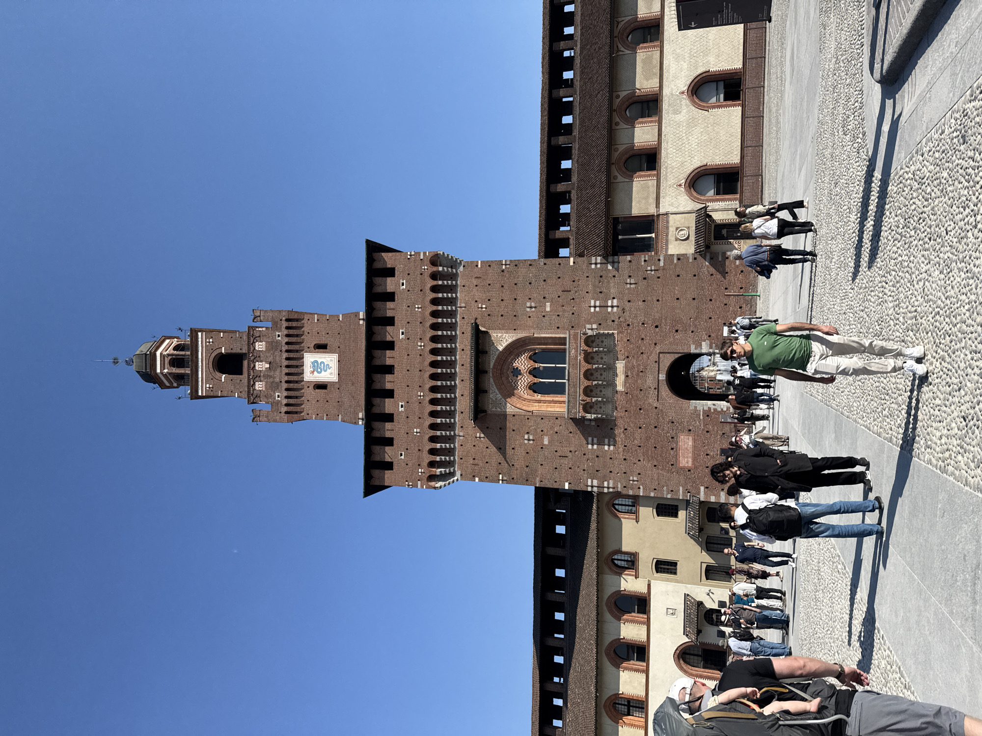 The image captures a bright, sunny view of the Sforza Castle (Castello Sforzesco) in Milan, Italy. The prominent Filarete Tower, a tall, red brick structure with a spired top, dominates the left side of the frame. The tower features a white clock face with black Roman numerals and distinctive decorative elements, including arched windows and crenellated battlements. A blue and white heraldic serpent symbol, representative of the Visconti/Sforza family, is visible on the tower's facade.
Adjacent to the tower, sections of the castle's brick walls and other buildings with lighter stone and arched windows are visible. In the foreground and midground, a large, paved courtyard is filled with numerous people, likely tourists or visitors, walking leisurely. The ground is a mix of small, textured pebbles or cobblestones and smoother flagstones.
The clear, vibrant blue sky suggests excellent weather. Bright sunlight illuminates the scene, casting long, sharp shadows from the people and buildings, indicating it is likely midday or early afternoon. People are casually dressed, engaged in sightseeing. One man in a green shirt and beige pants stands prominently in the mid-right. Another person in the foreground wears a baby carrier. A small, black sign with white text is affixed to a building but is too distant to read. The overall scene depicts a bustling public space at a historical landmark on a clear day.