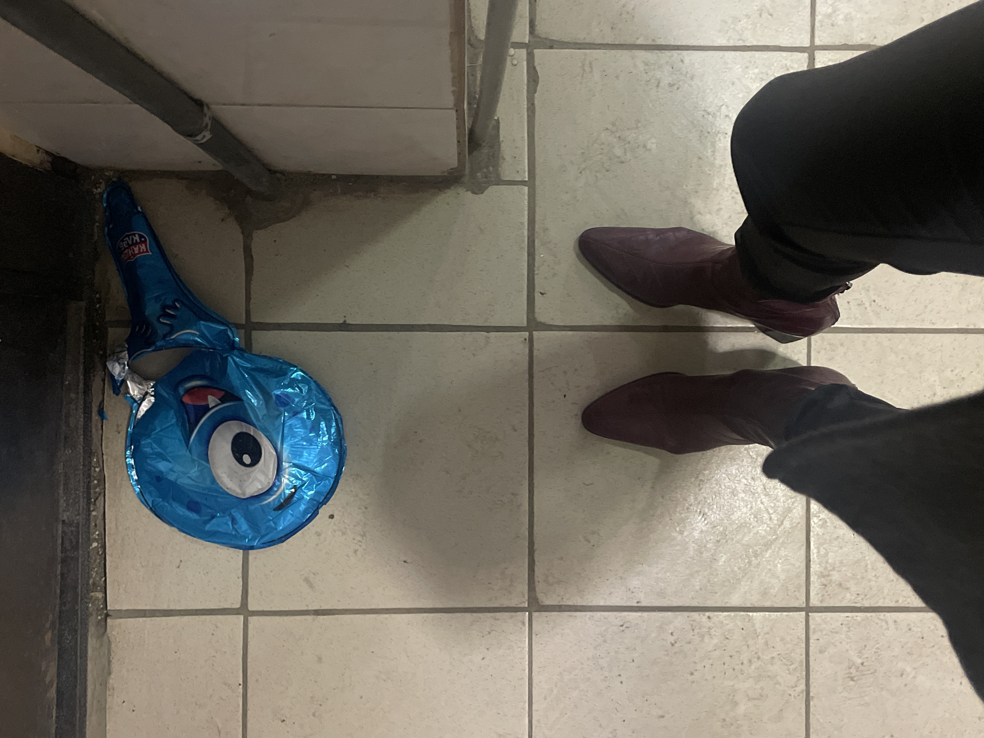 The image depicts a tiled floor with a deflated blue balloon featuring a cartoonish eye design near a wall. Next to the balloon are two legs clad in dark pants and maroon-colored pointed boots. The location appears to be indoors, likely a bathroom or similar space with tiled flooring. There is no information to confirm the location is Moscow, Russia.